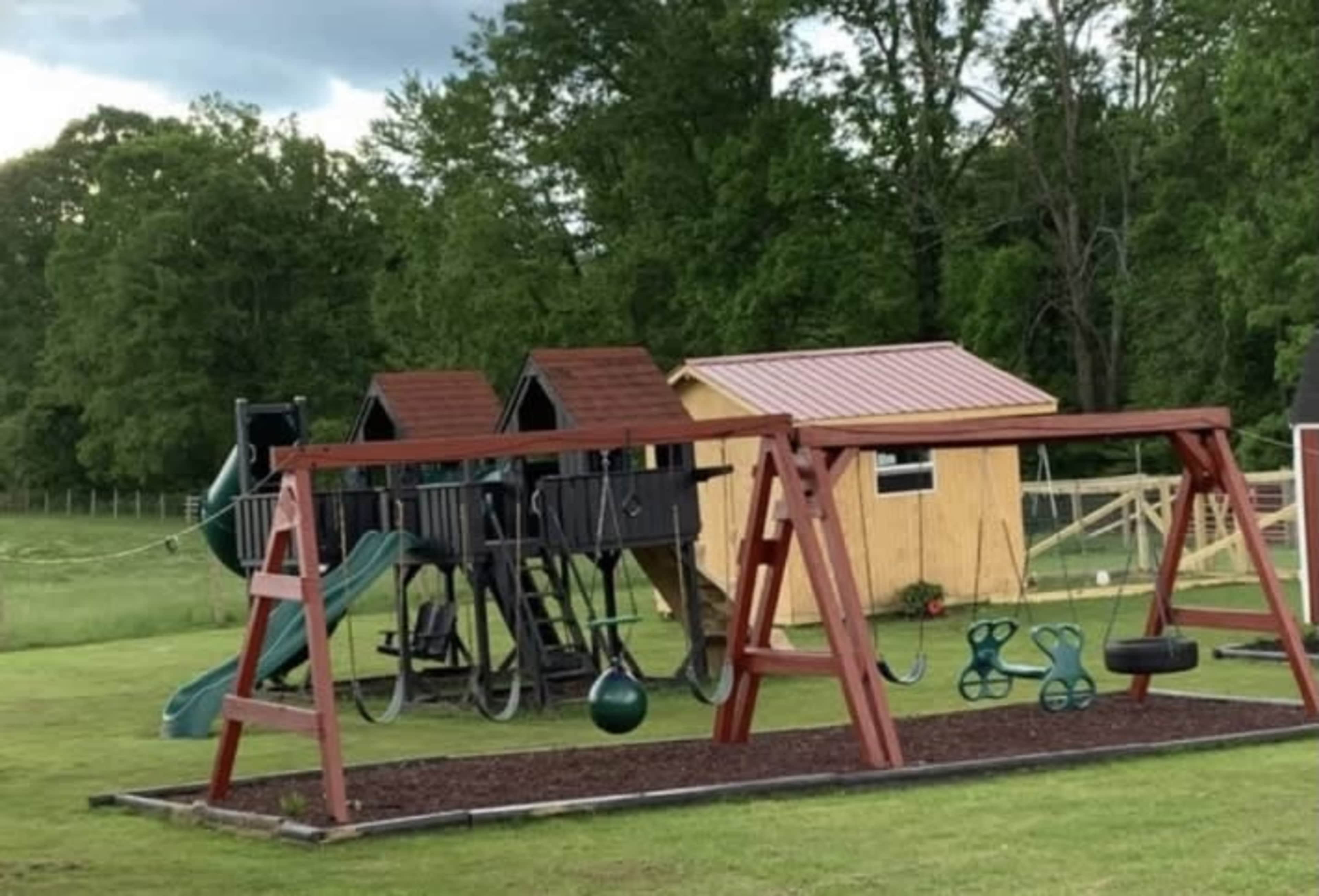 The image shows a playground with a wooden swing set, a slide, and two small playhouses, all set in a grassy area surrounded by trees.