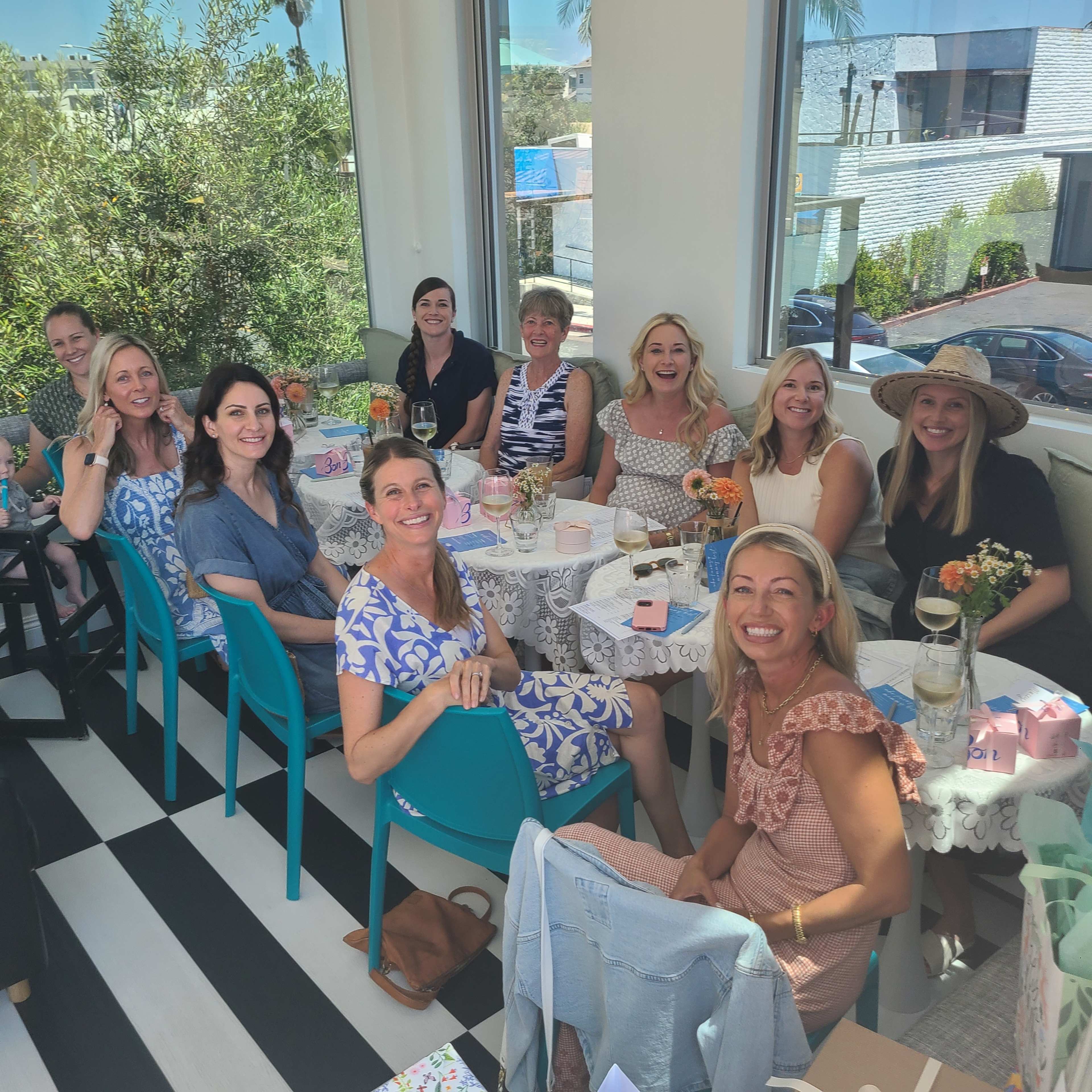 A group of women is seated at tables in a bright, modern café, enjoying drinks and a meal together.