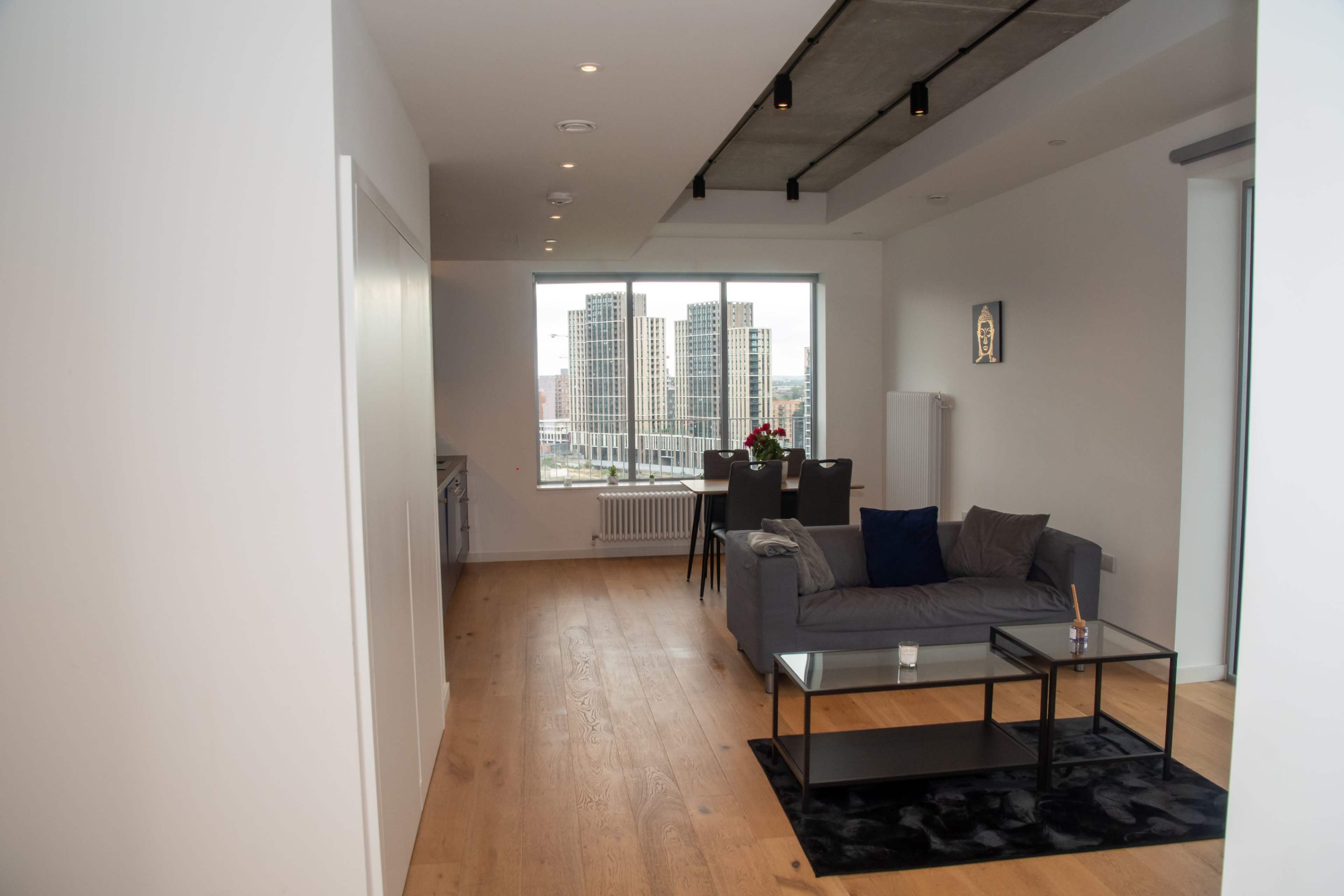 A modern apartment interior with a living area featuring a sofa, coffee table, dining table, and large windows overlooking a cityscape.