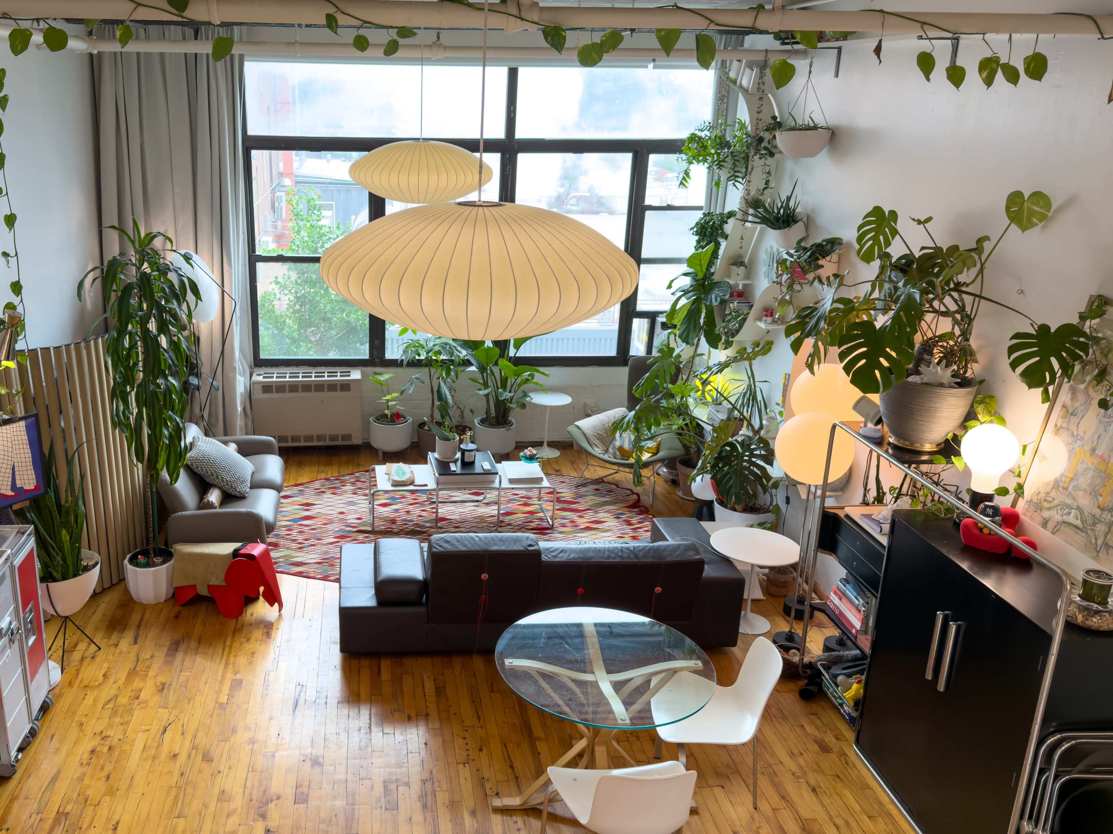 The image shows a bright, open living space with green plants, a large light fixture, a circular glass table, and a dark sofa, framed by large windows.