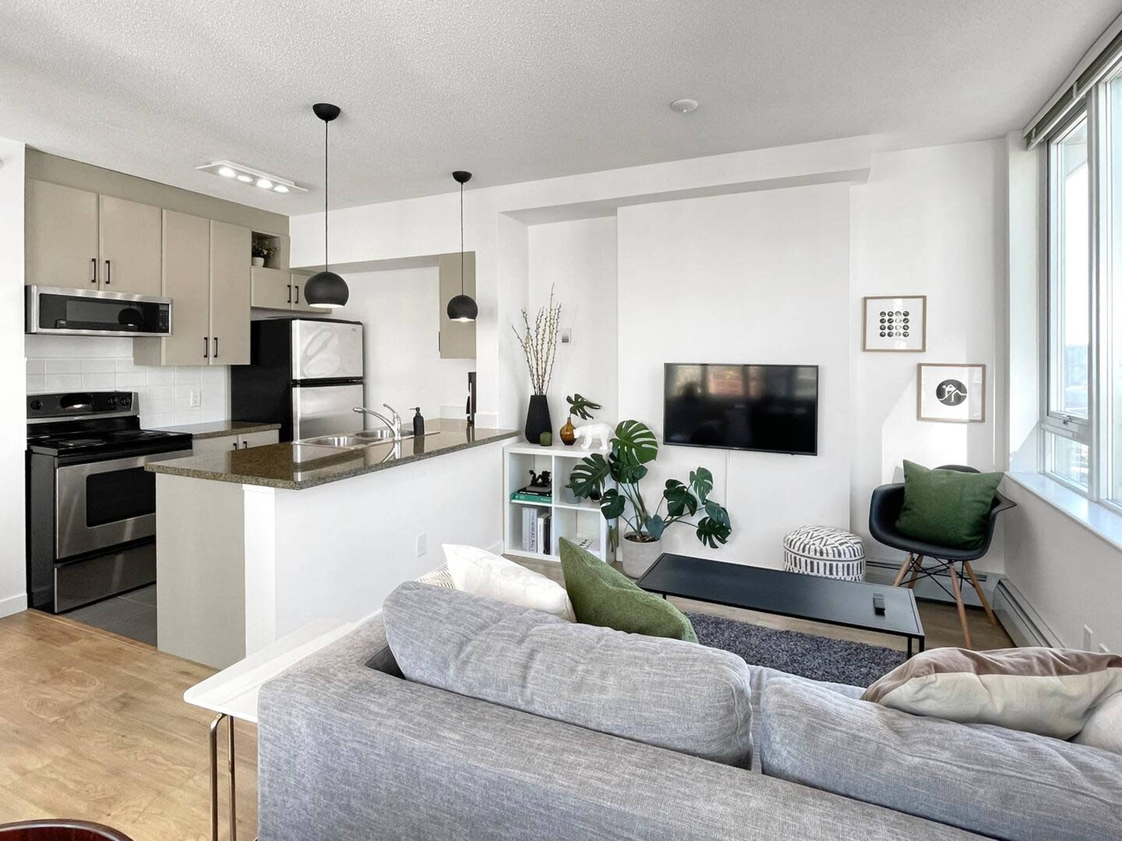 The image shows a modern, open-concept living area with a kitchen featuring sleek cabinets, a small dining space, and a cozy seating area with a gray sofa and a mounted television.