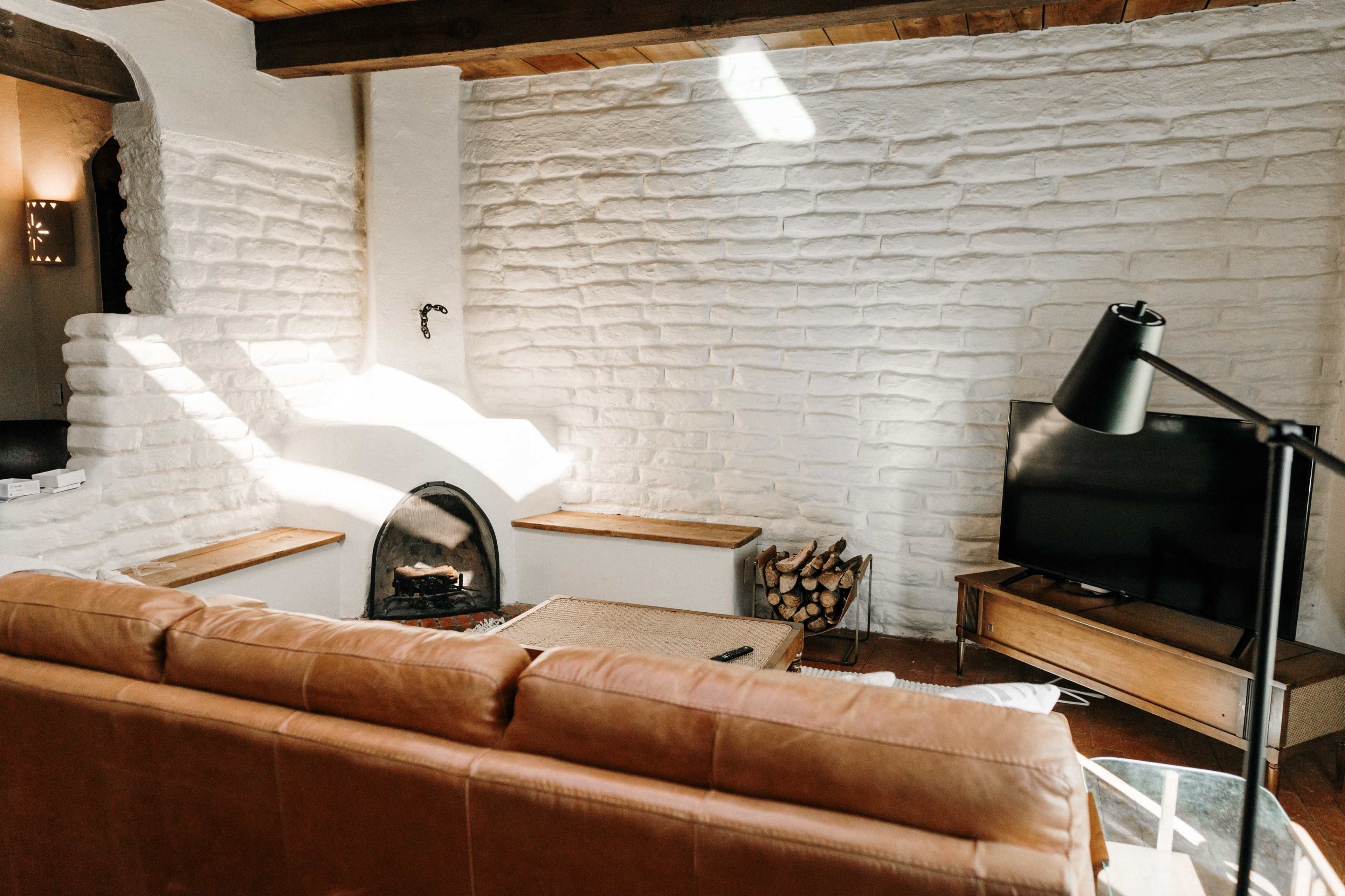 A cozy living room with a white stone wall, a brown leather sofa, a modern lamp, and a television, complemented by a fireplace and stacked firewood.