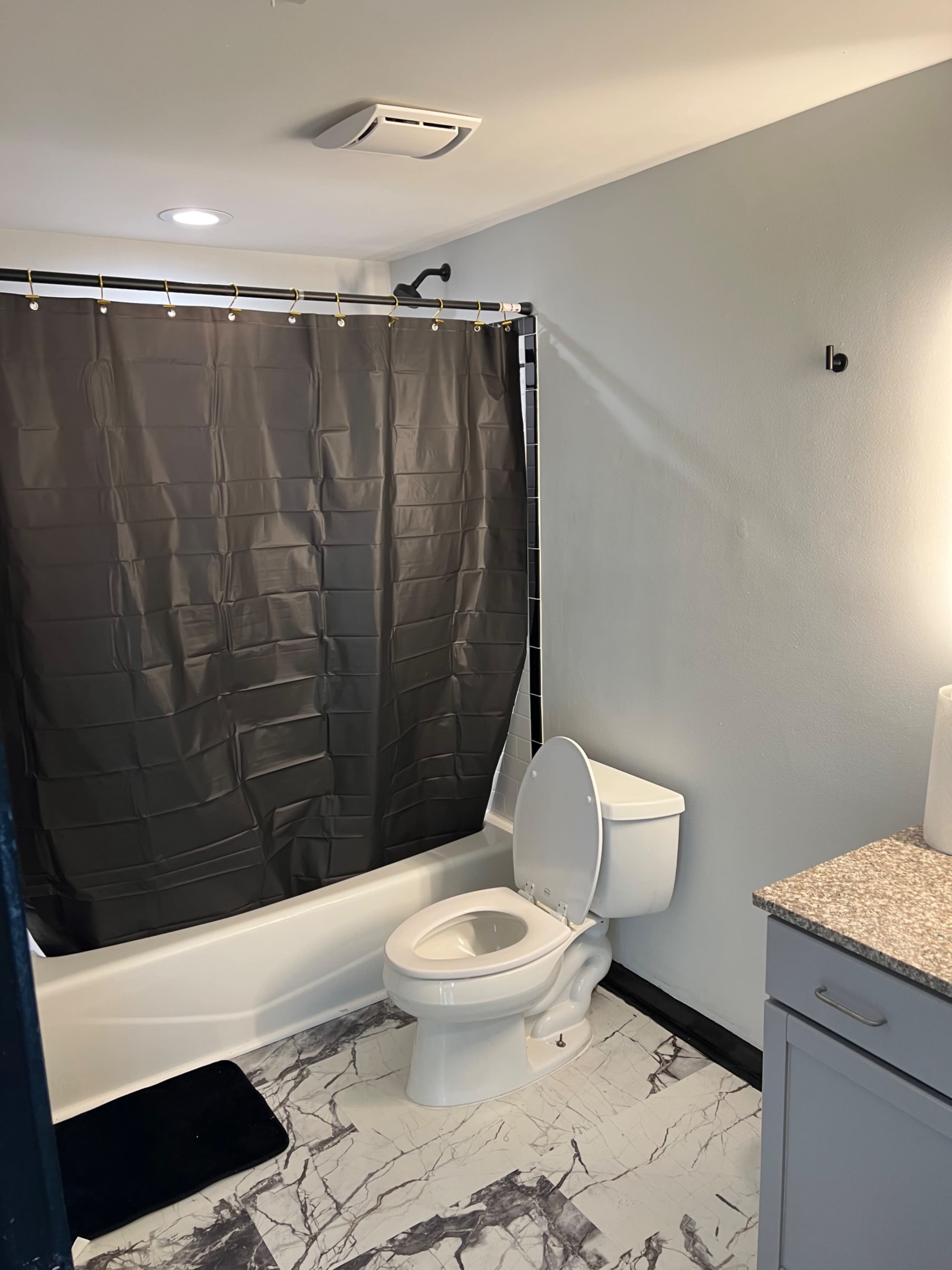 The image shows a bathroom featuring a black shower curtain, a toilet, and a sink with a granite countertop, all set against a gray wall and a marble-patterned floor.