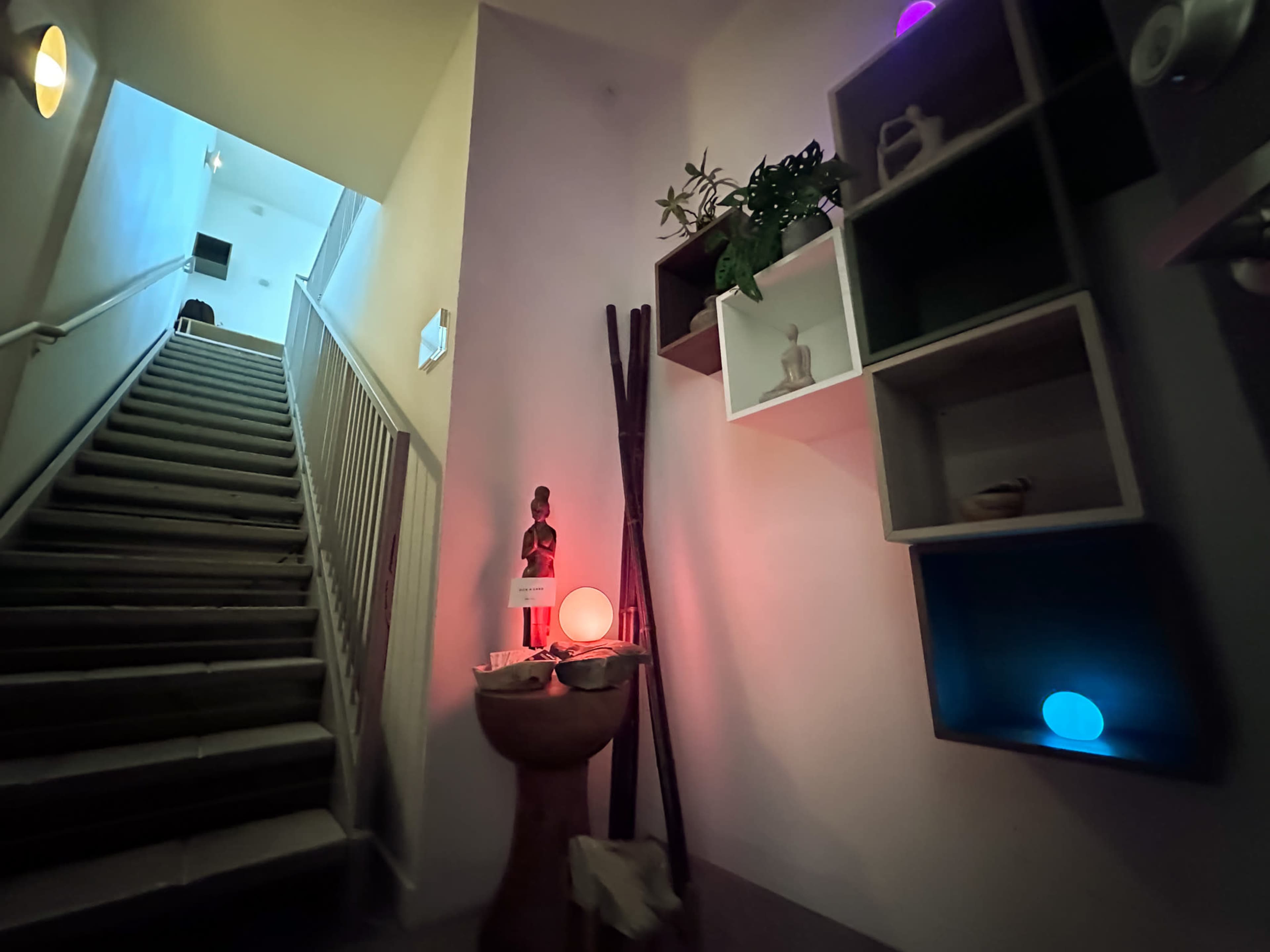 A staircase leads upward beside a wall adorned with shelves containing decor, including a statue and colored lights.