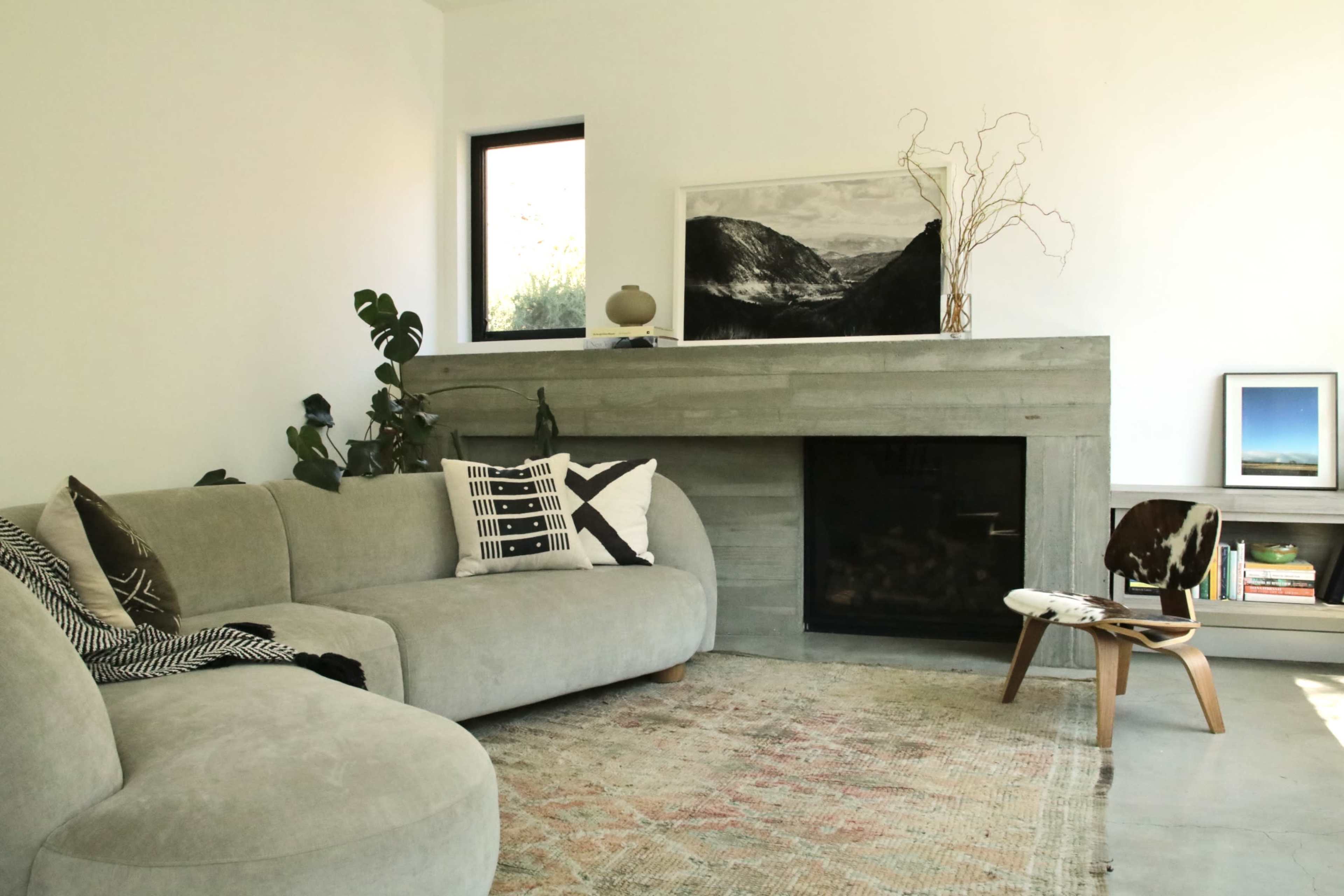 The scene shows a modern living room featuring a curved gray couch, a concrete fireplace with artwork above it, and decorative elements like plants and a small side table.