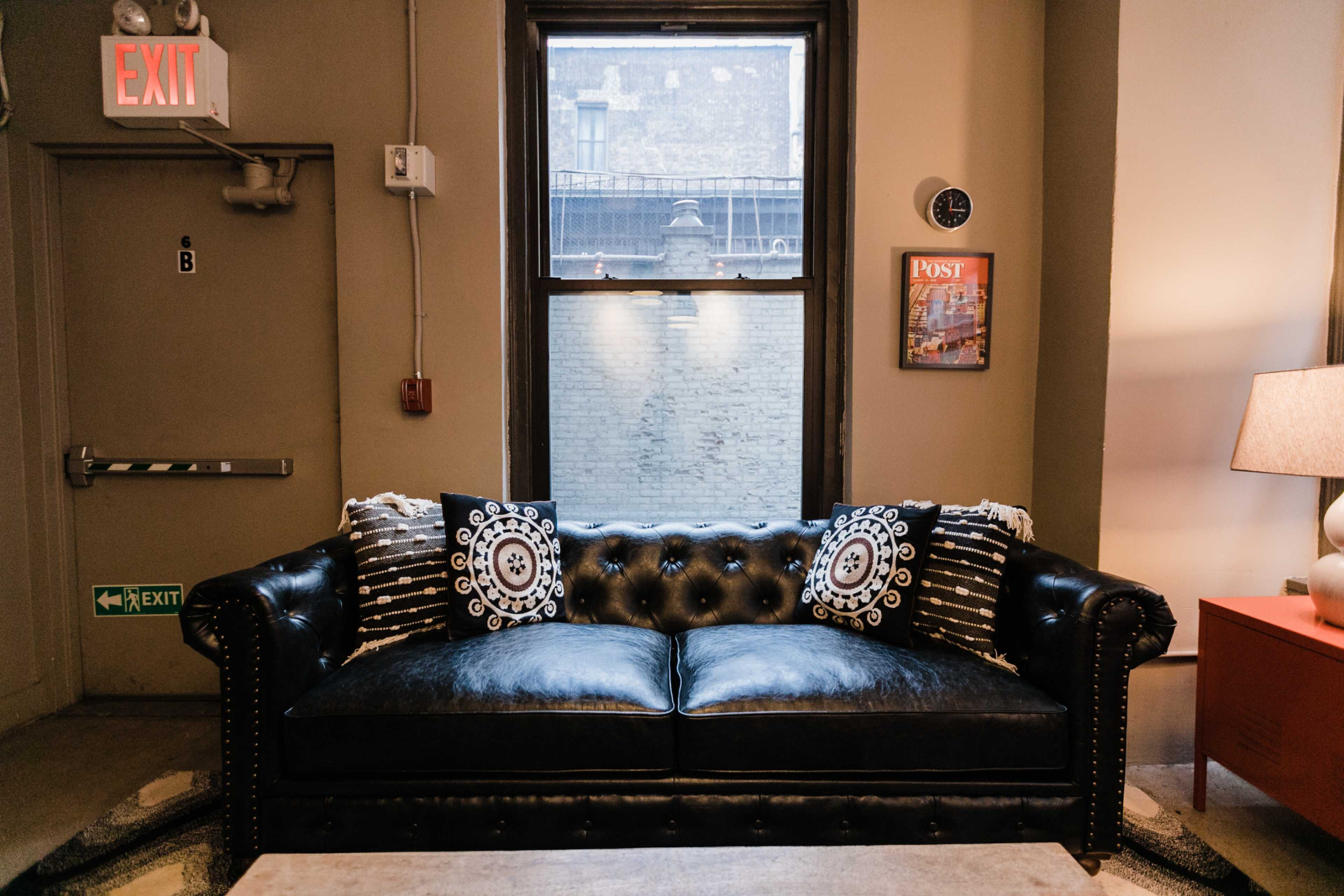A black tufted leather sofa with decorative pillows is positioned against a wall beneath a window in a room with neutral decor.