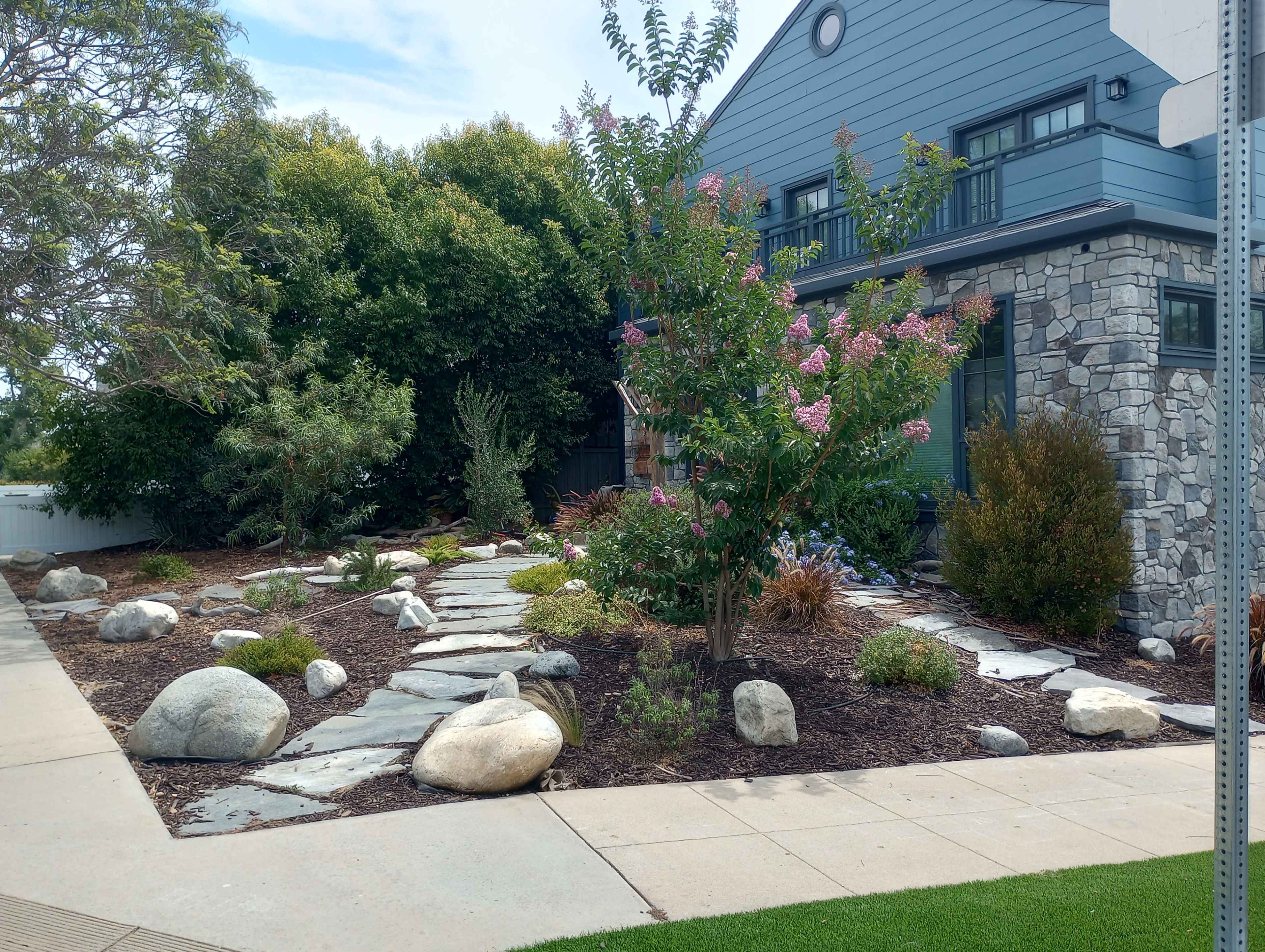 A garden features stone pathways, various plants, and decorative rocks alongside a modern house.