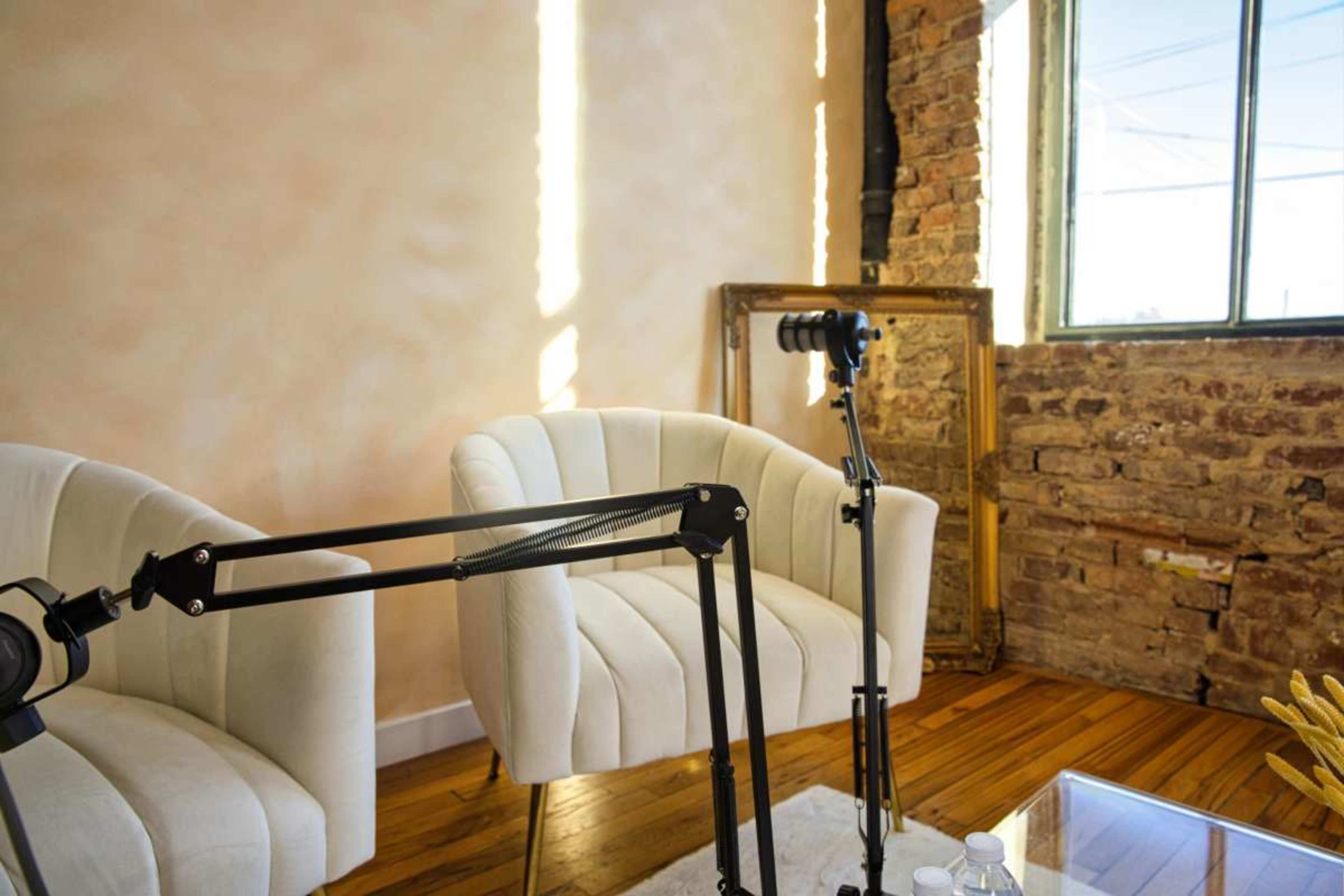 The image shows a cozy interior space featuring two white armchairs, a large window, and a camera setup on a tripod, with exposed brick walls and wooden flooring.