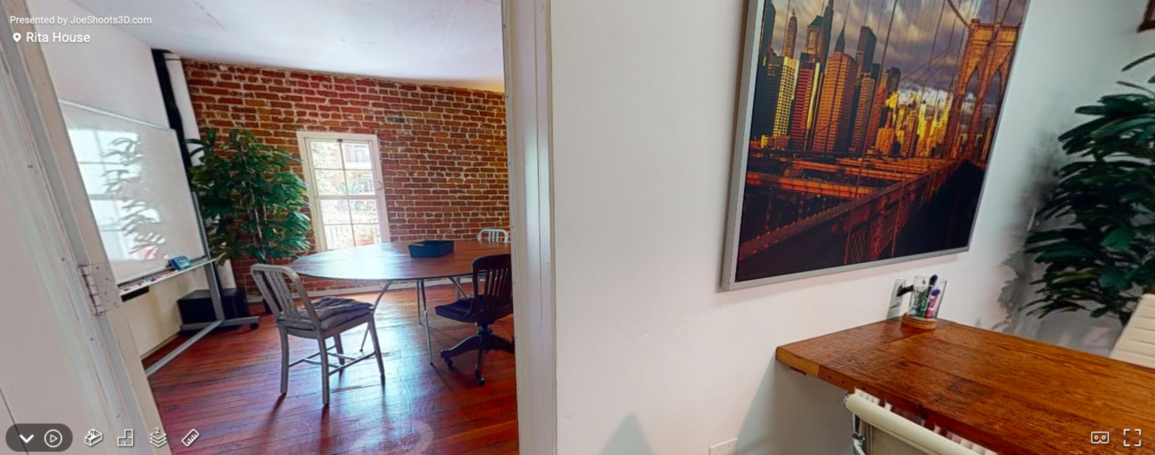 A bright room with exposed brick walls, containing a large table and chairs, connected to another area featuring a desk, a plant, and a large wall photo of a city skyline.