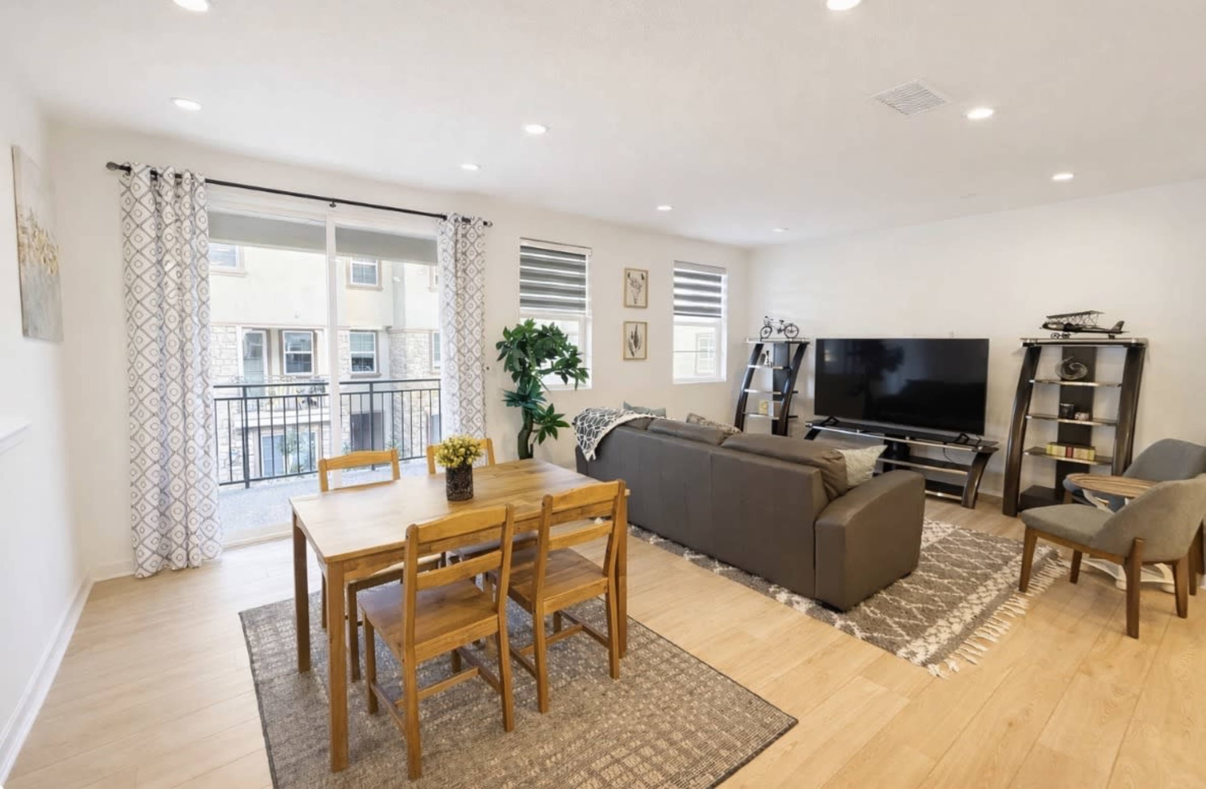 The image shows a modern living room with a dining area, featuring a sofa, a television stand, a table with chairs, and large windows allowing natural light to enter.