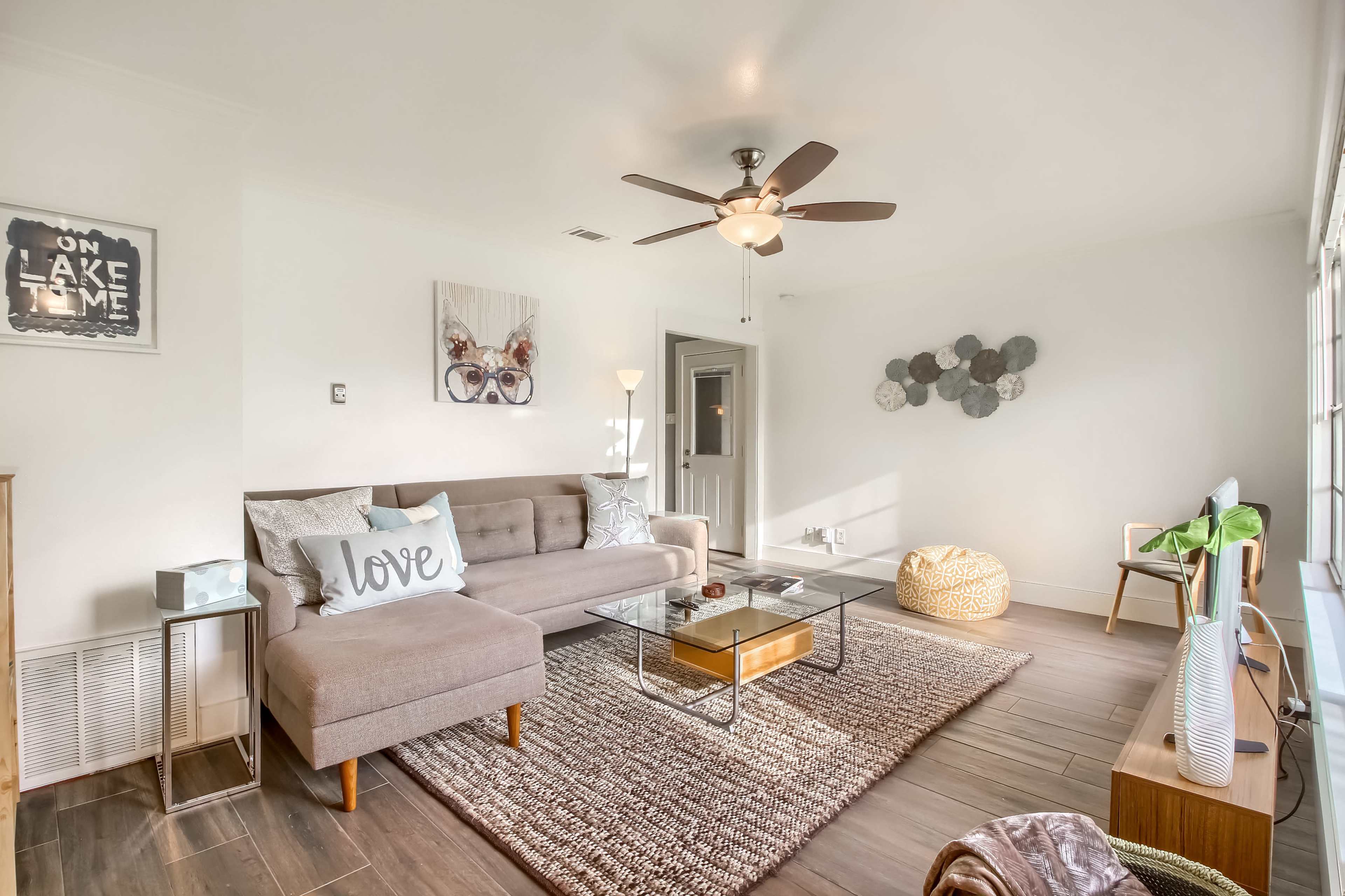 A bright and airy living room features a gray sofa, a coffee table, a ceiling fan, and decorative wall art.