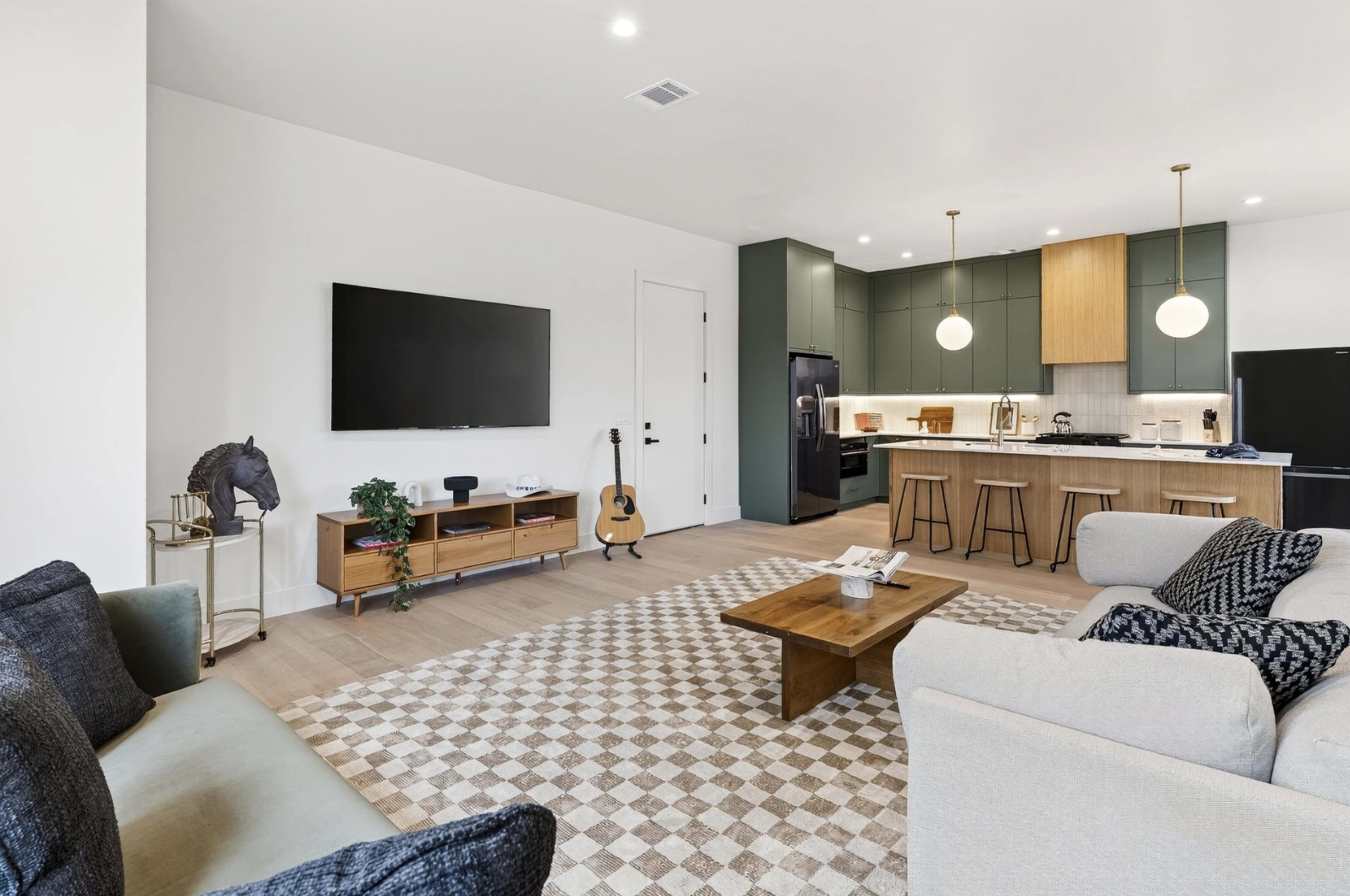 The image shows a modern living room with a television mounted on the wall, a coffee table in the center, and an open kitchen area featuring green cabinetry and pendant lights.