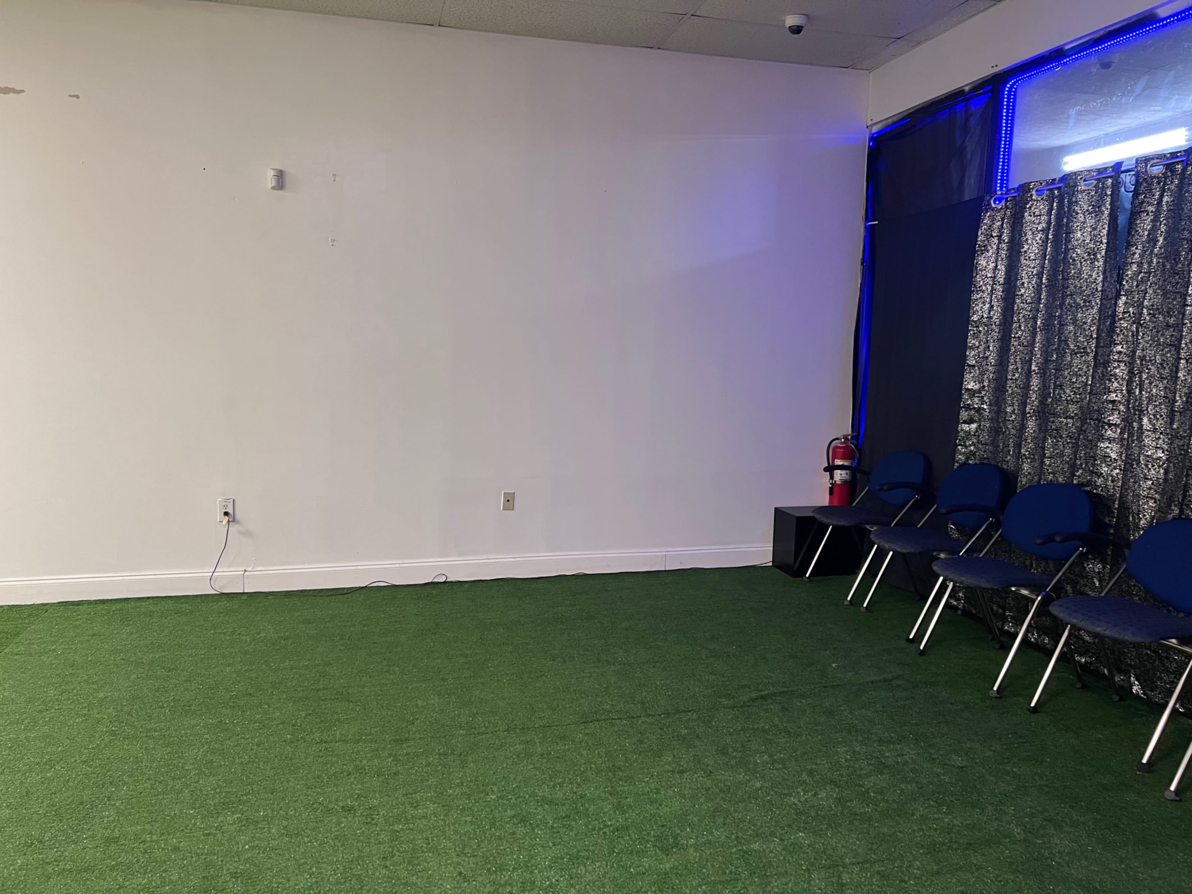 An empty room with green artificial turf, a row of blue chairs, and a fire extinguisher against the wall.