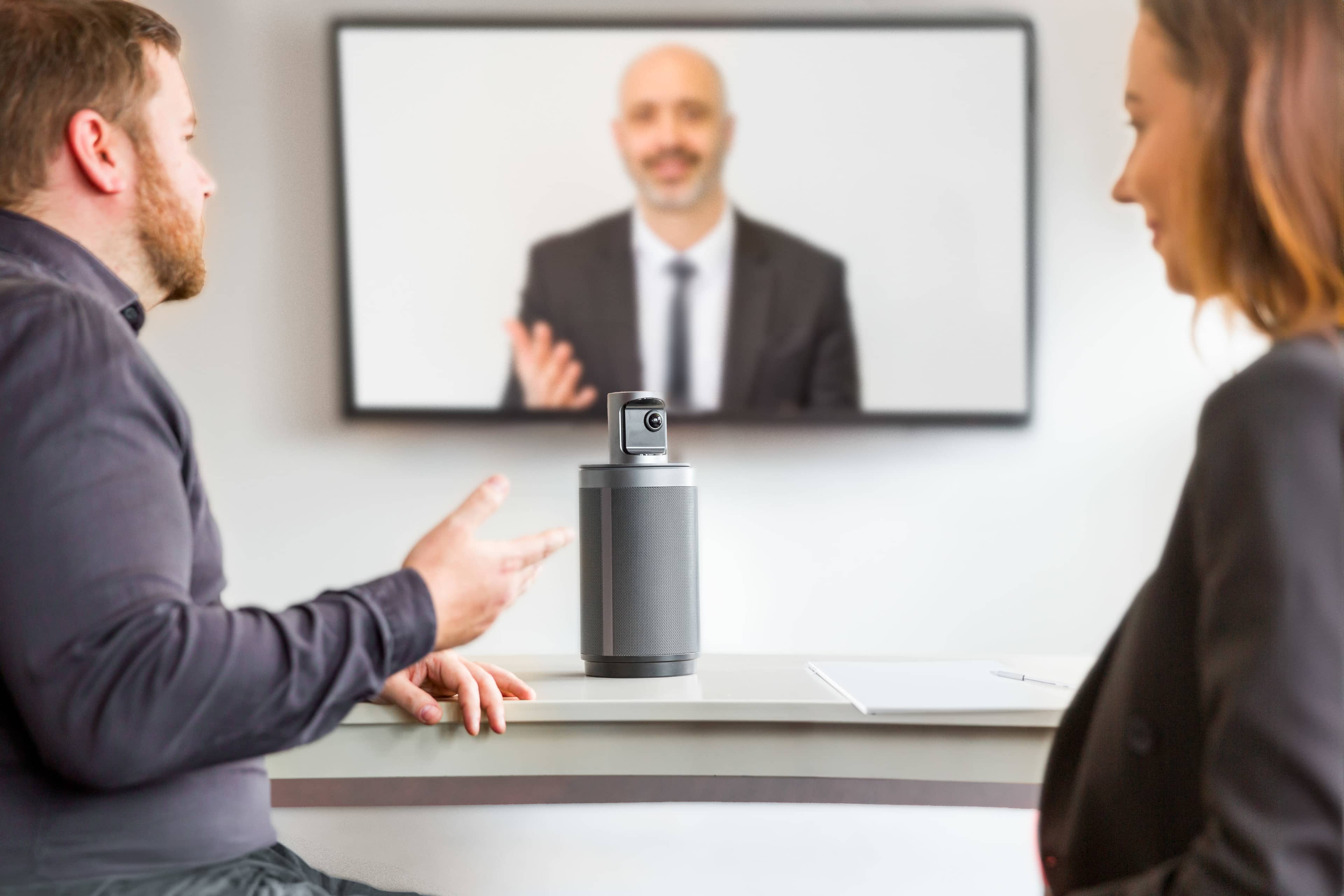 A man and woman engage in a video conference at a table with a speaker device in the center, while a third person appears on a screen in the background.