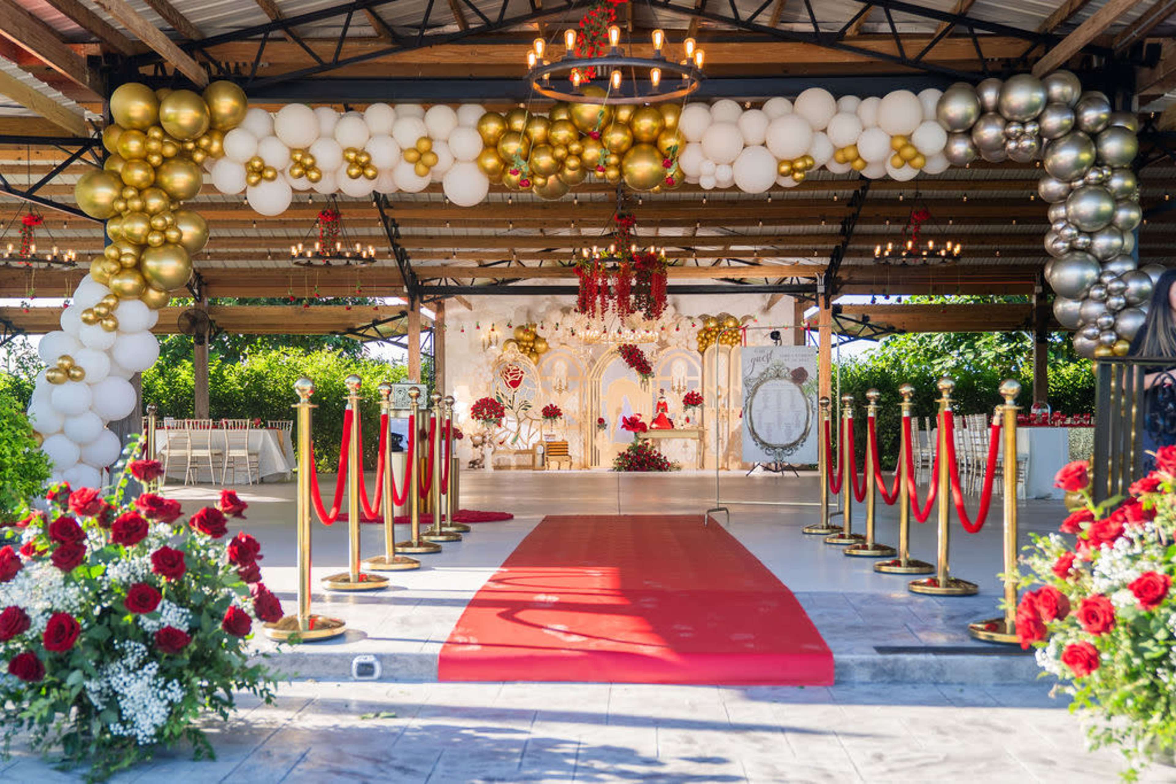 The image shows a decorated entrance featuring a red carpet, gold and white balloon arches, floral arrangements, and a backdrop with ornate decor set for a wedding celebration.