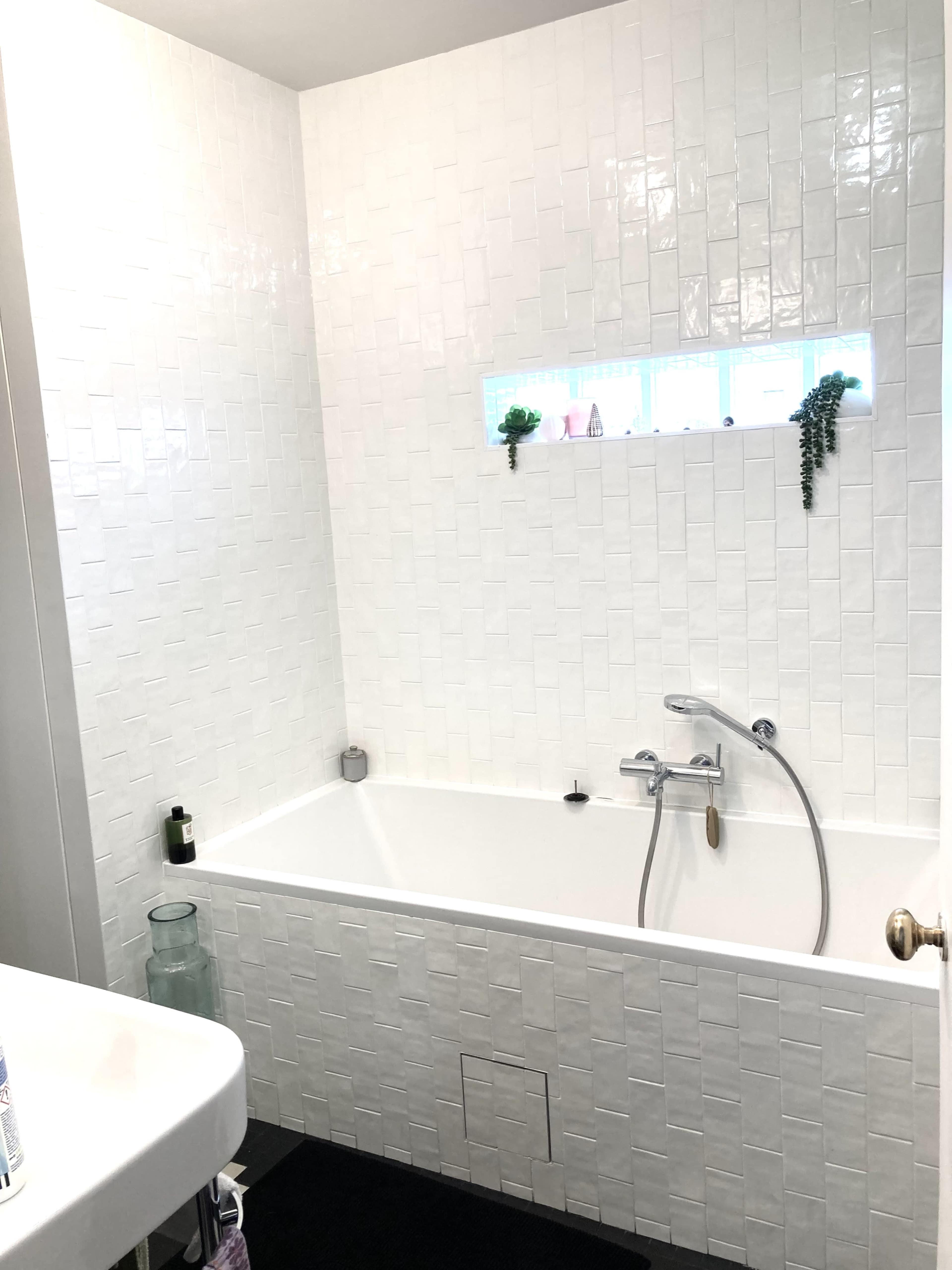 A modern bathroom featuring a white tiled wall and a bathtub with a shower fixture, complemented by a small window with decorative plants.