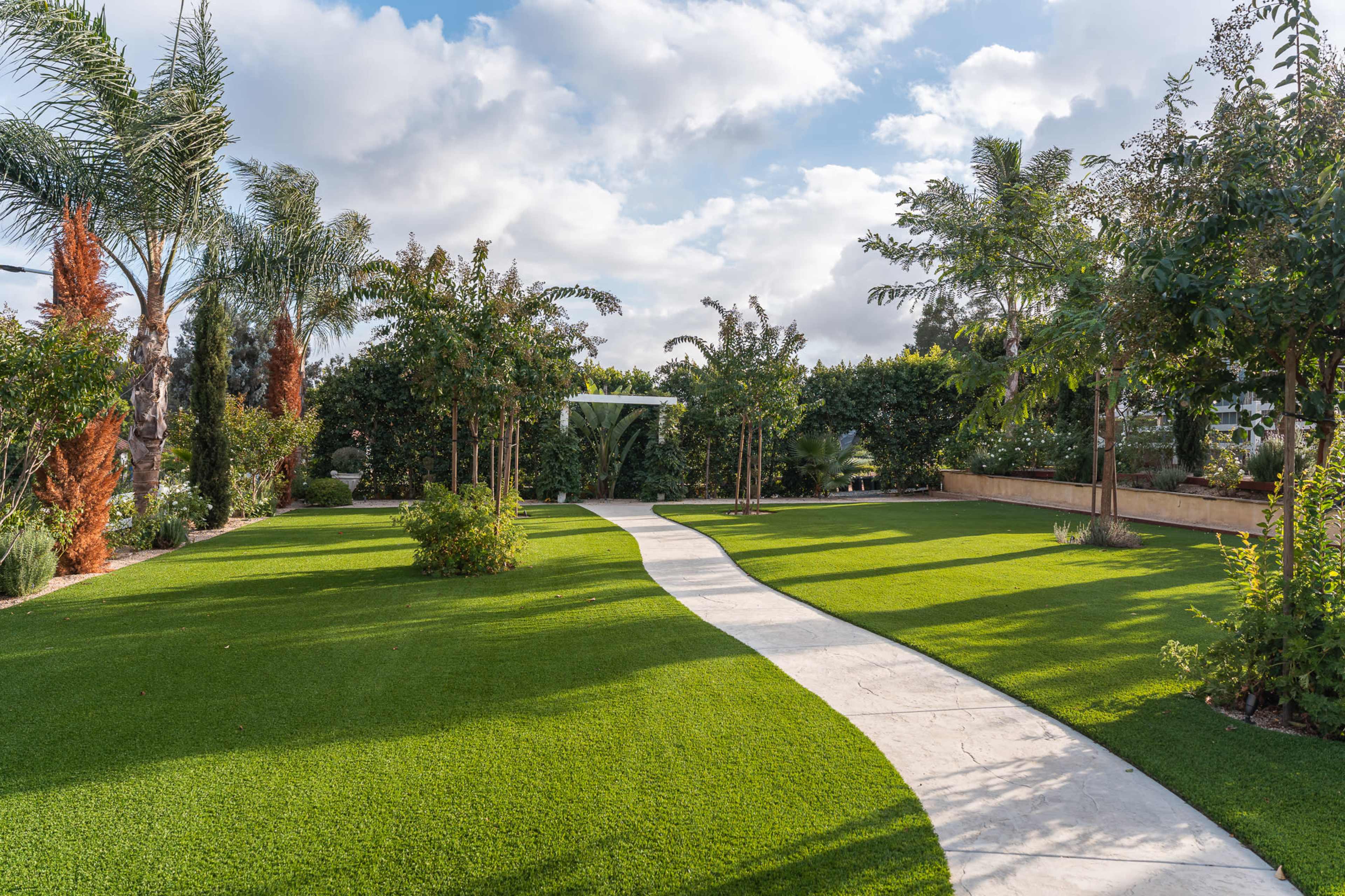 A landscaped garden features a manicured lawn with a curved stone pathway surrounded by various trees and shrubs under a partly cloudy sky.
