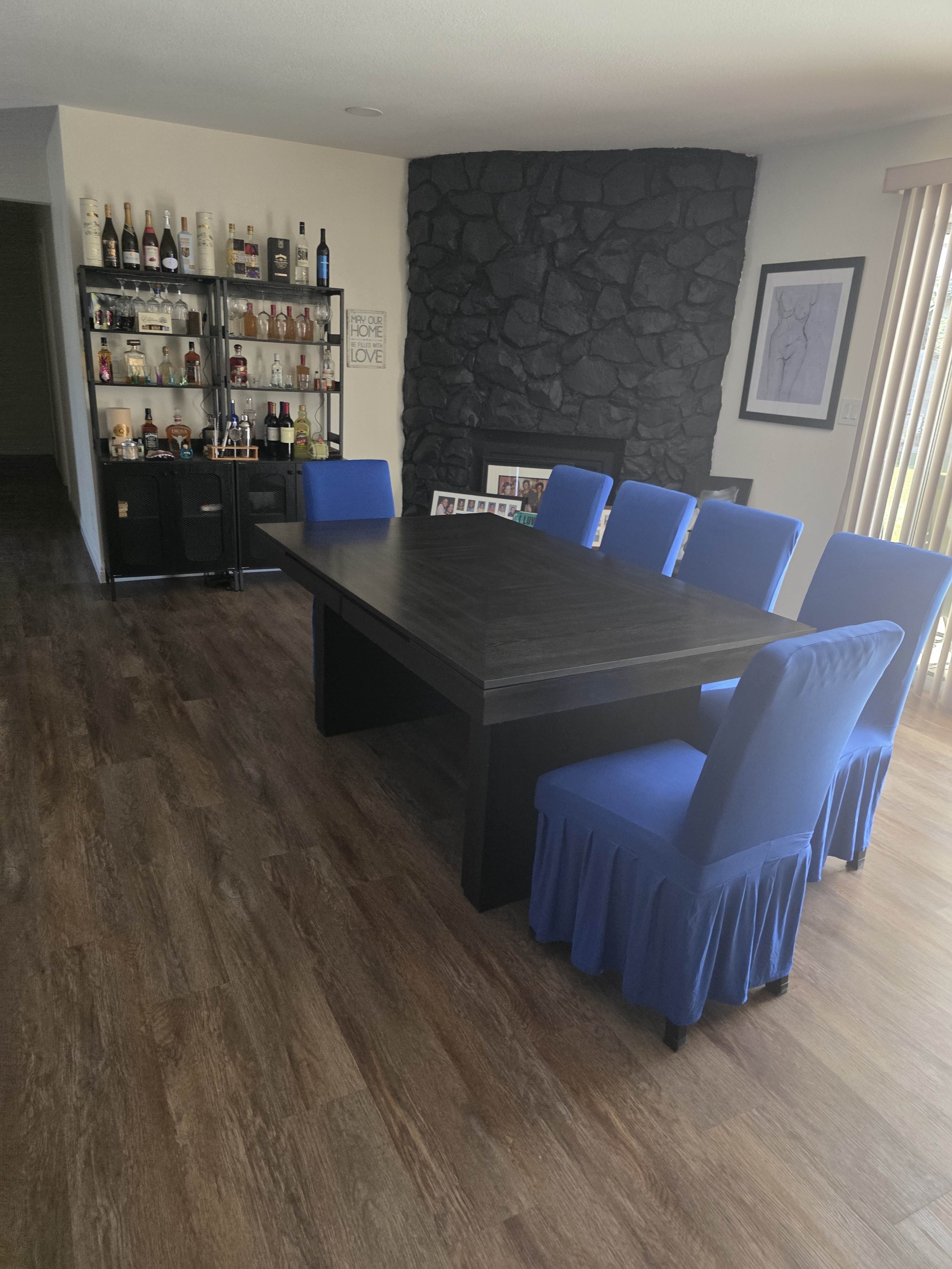 The image shows a dining area with a large rectangular table surrounded by six blue chairs, alongside a stone wall and a shelving unit displaying various bottles.