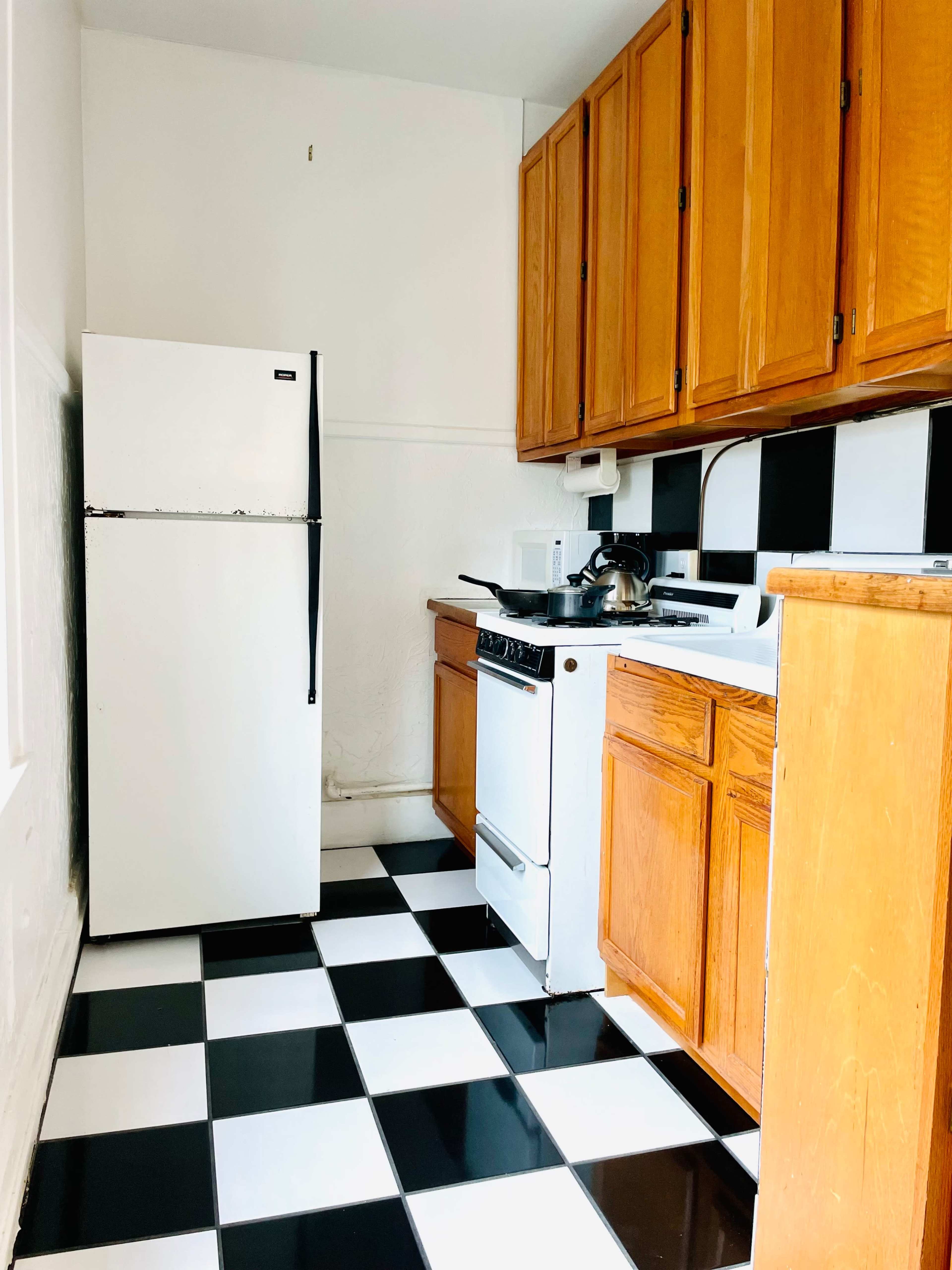 A small kitchen features black and white checkered flooring, wooden cabinets, a refrigerator, and a stove.