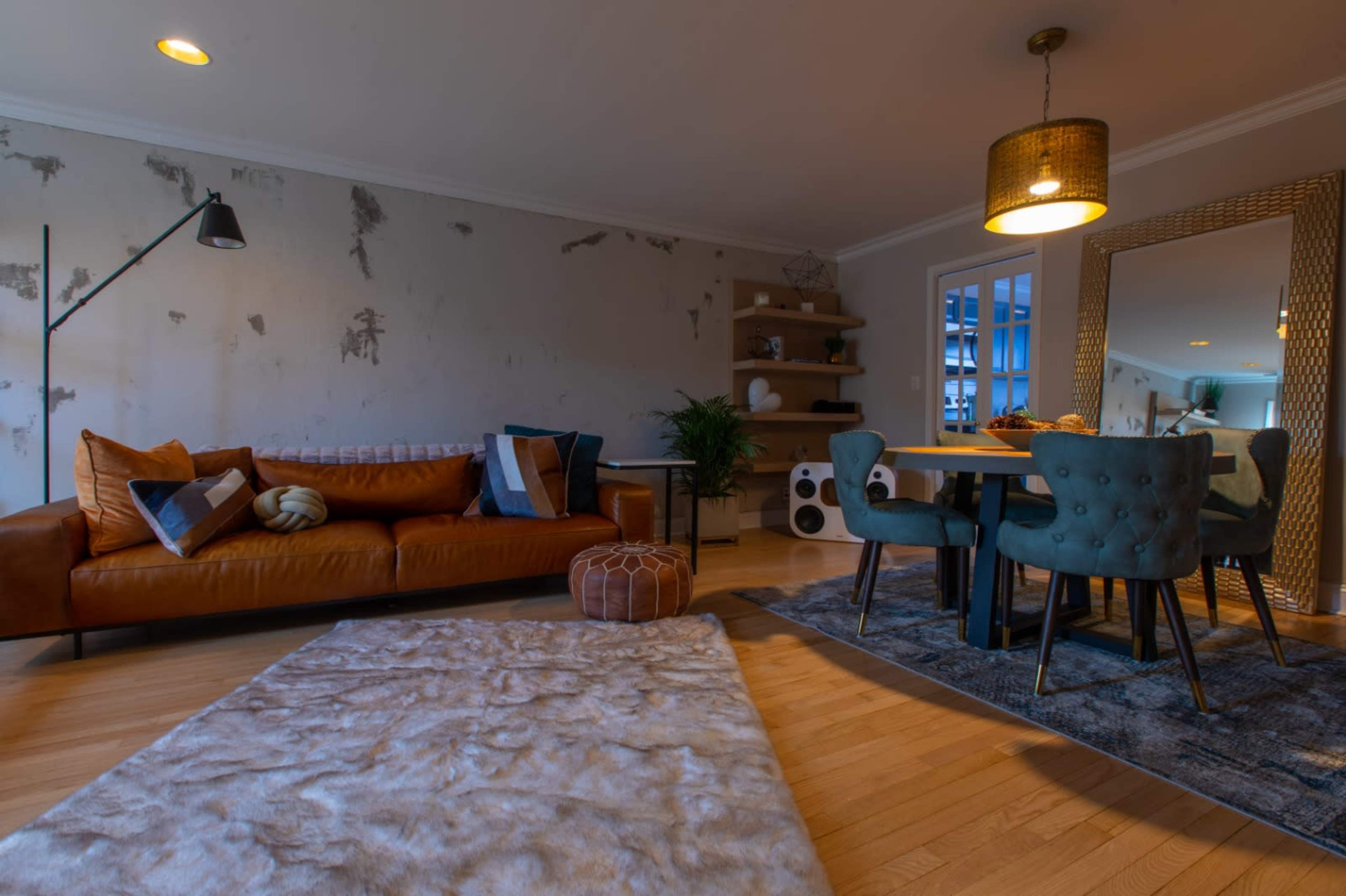 A living area featuring a brown leather sofa, a round dining table with upholstered chairs, and a large mirror on the wall, all set against a partially unfinished wall.