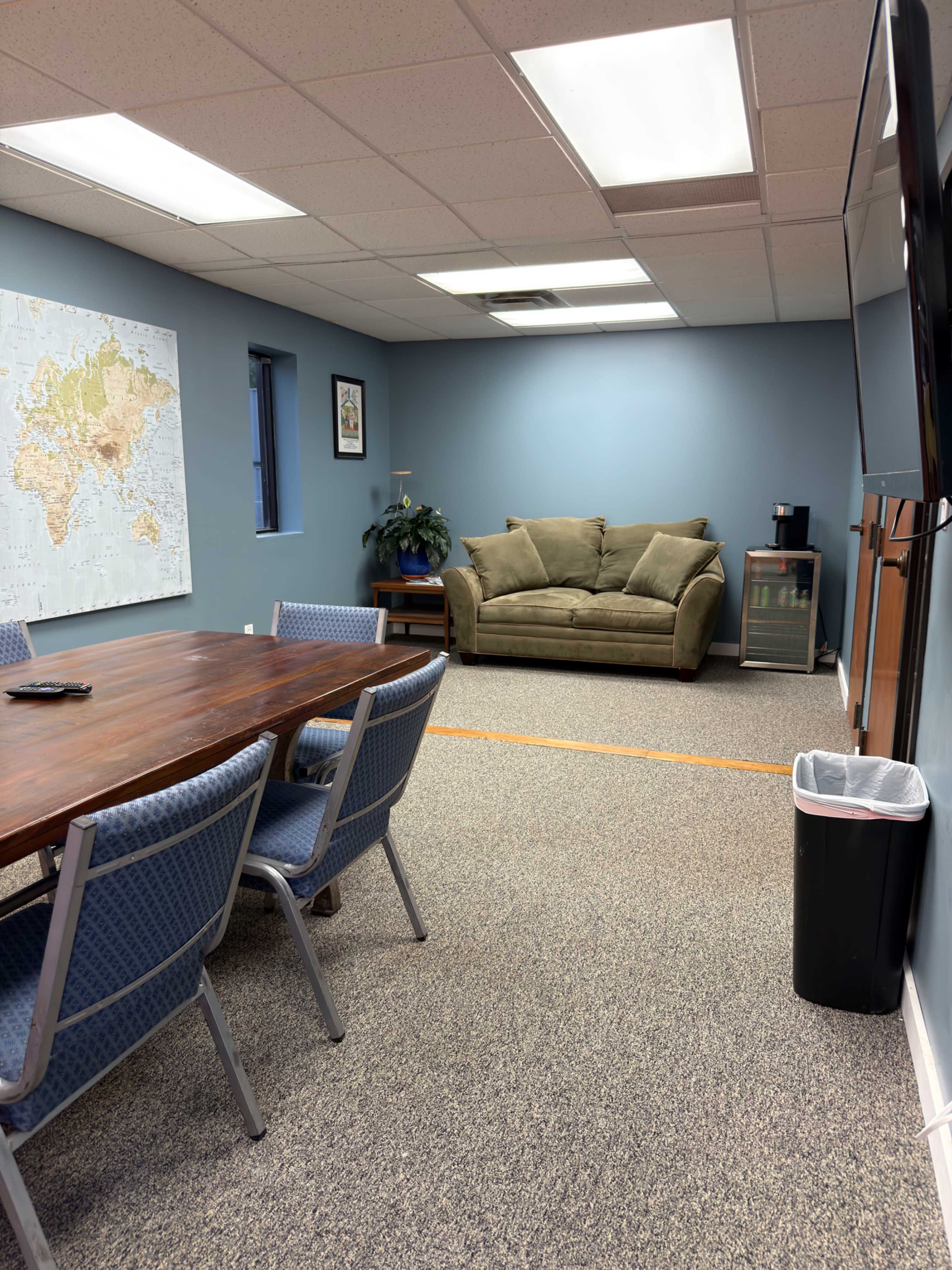 The image shows a meeting room with a large wooden table, several chairs, a couch against the wall, a potted plant, and a map on the wall.