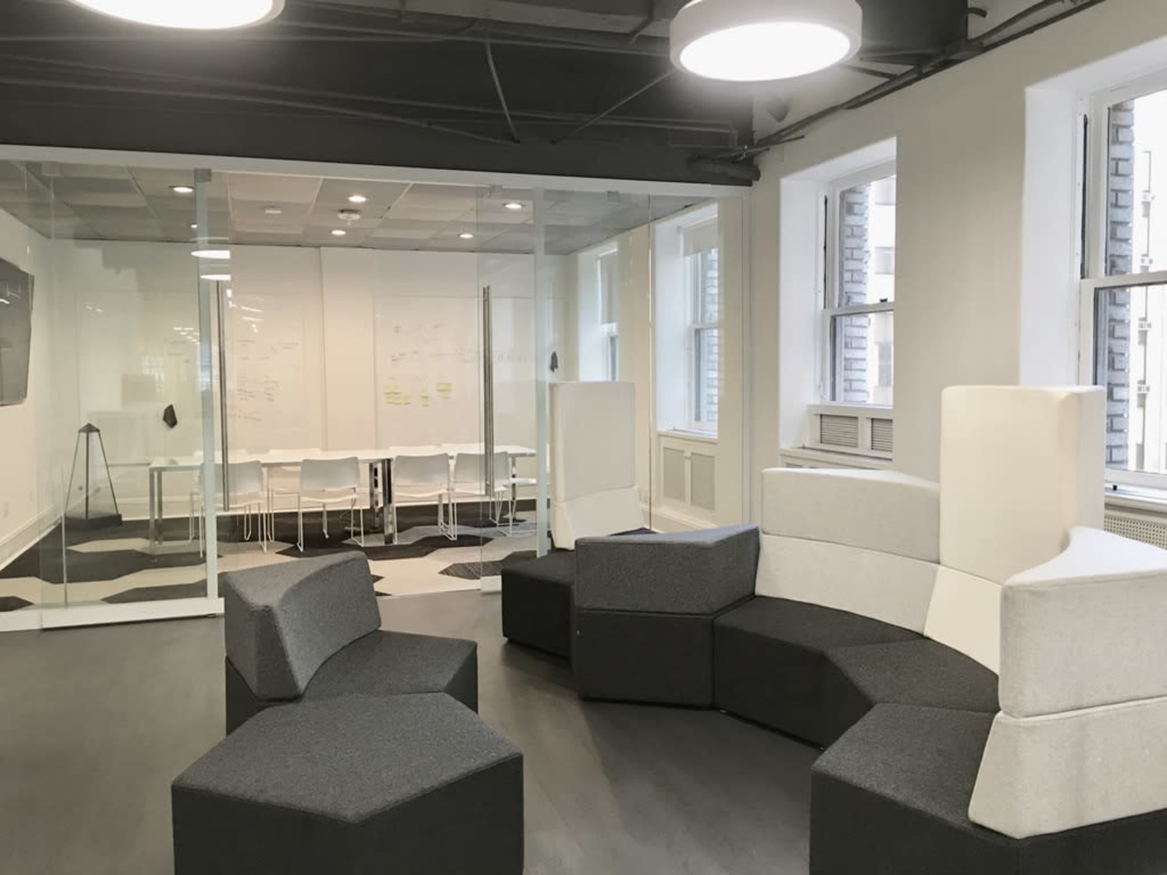 A modern office space featuring a seating area with modular gray furniture, adjacent to a glass-walled meeting room equipped with a table and chairs.