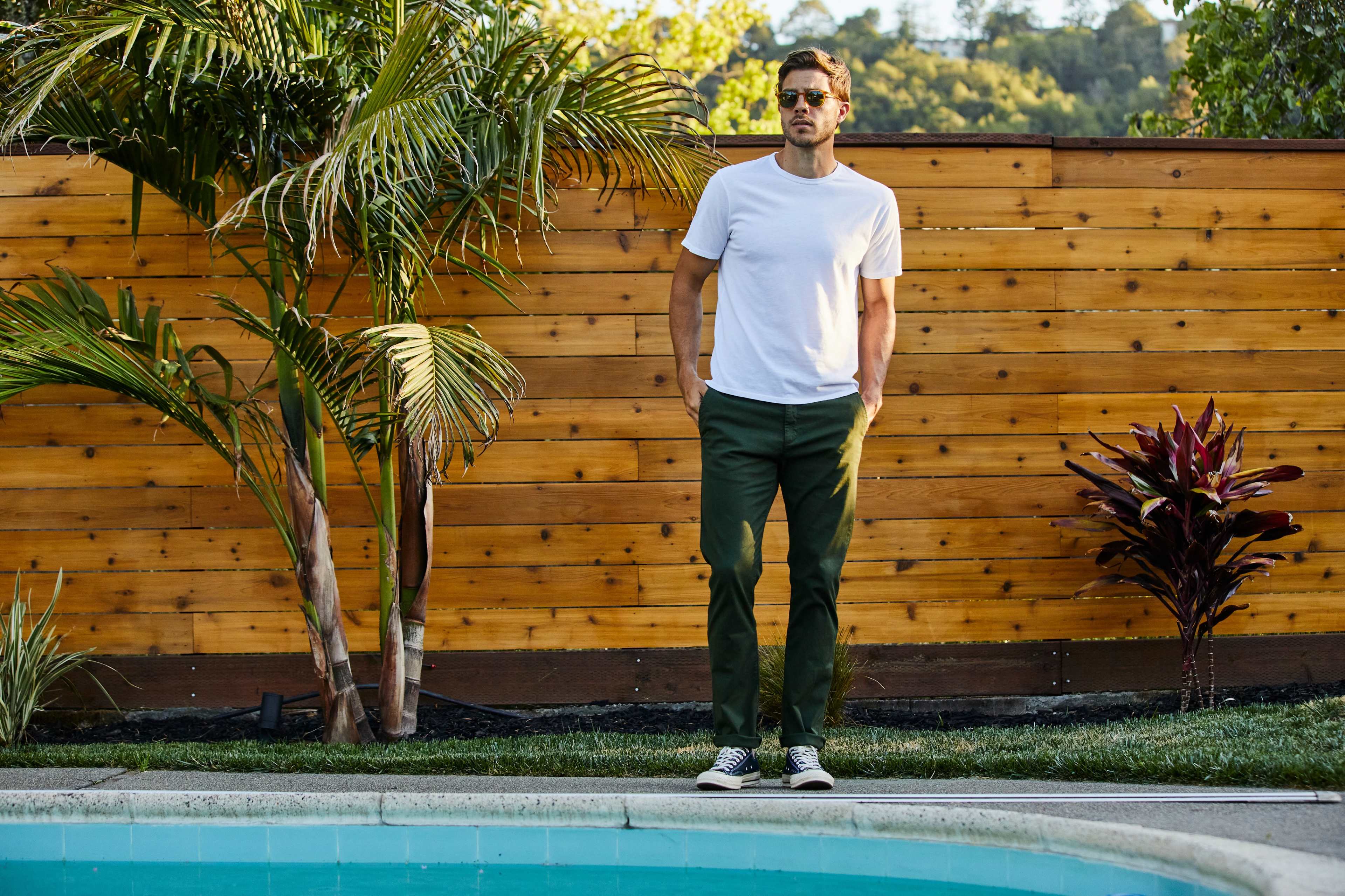 A man in a white t-shirt and green pants stands by a swimming pool, surrounded by palm trees and a wooden fence.