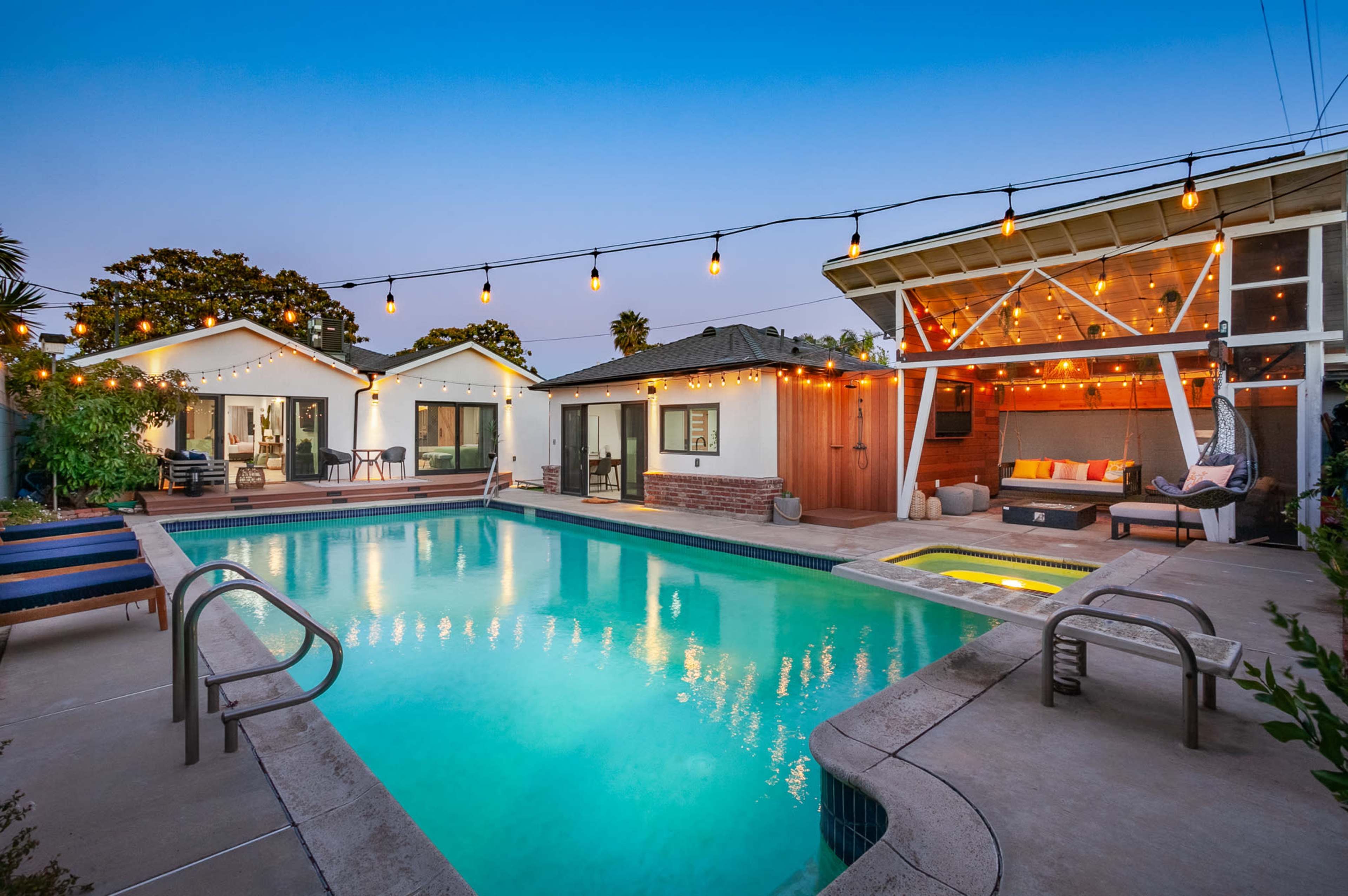 A well-lit backyard features a swimming pool surrounded by modern seating areas and a covered patio with string lights.