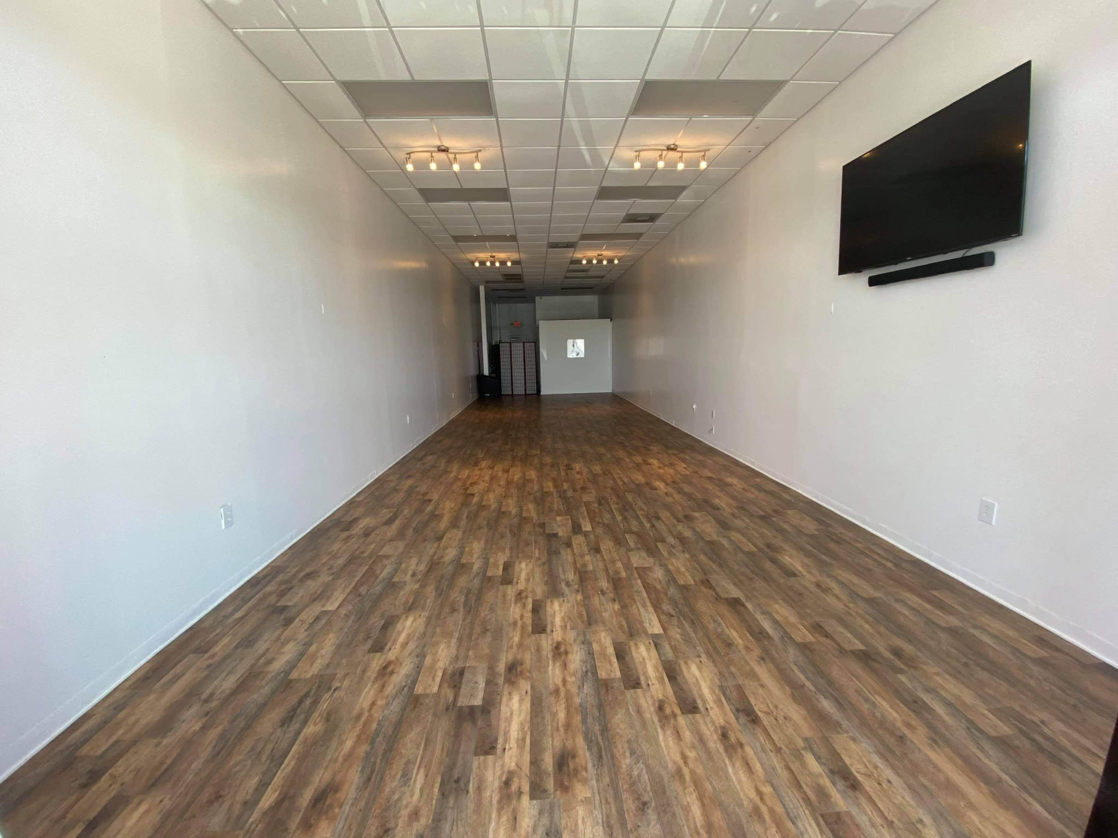 A spacious, empty storefront with wooden flooring and bright overhead lights.