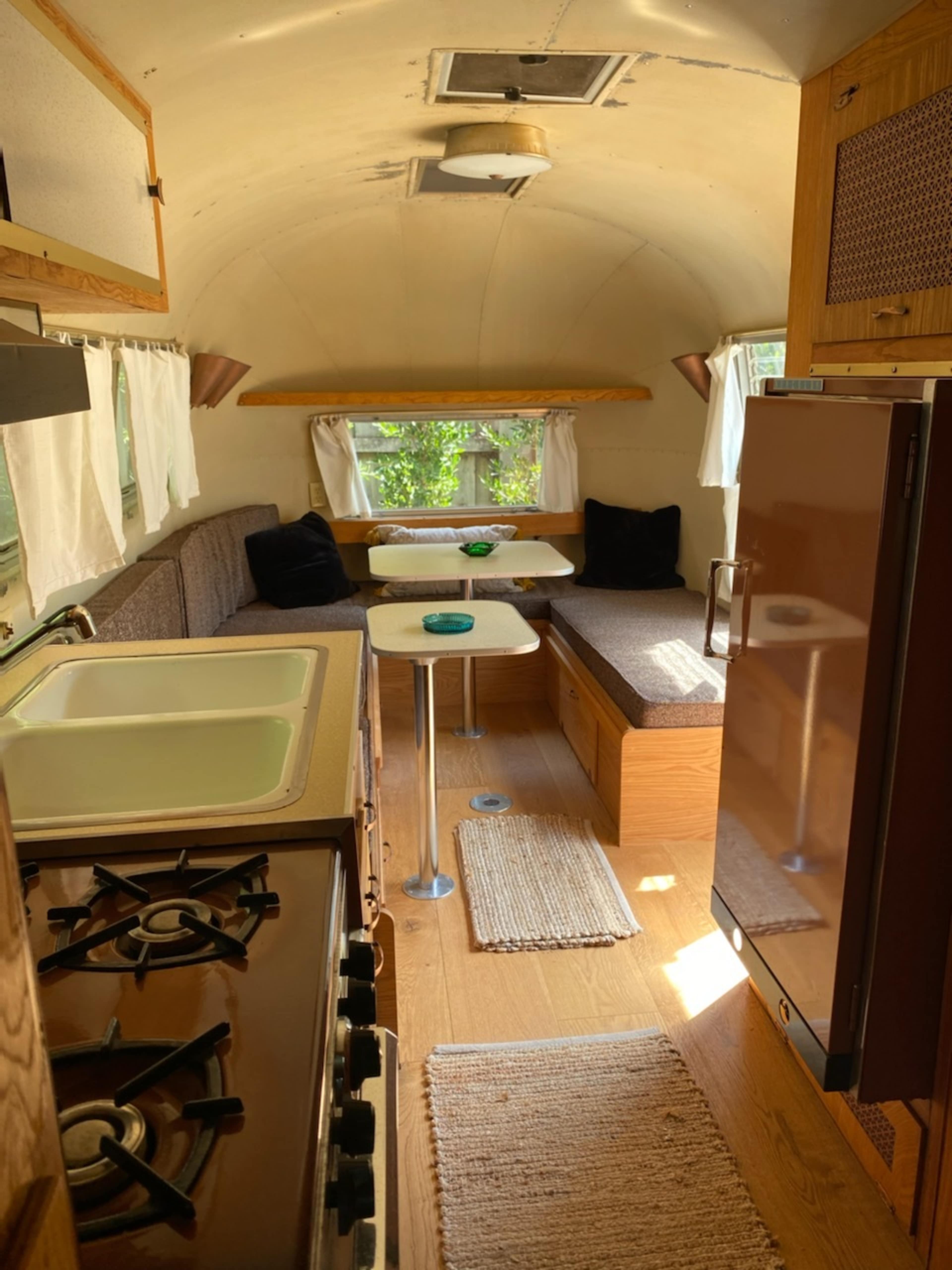 The interior of a vintage trailer features a kitchenette with a stove, seating area with a table, and large windows.