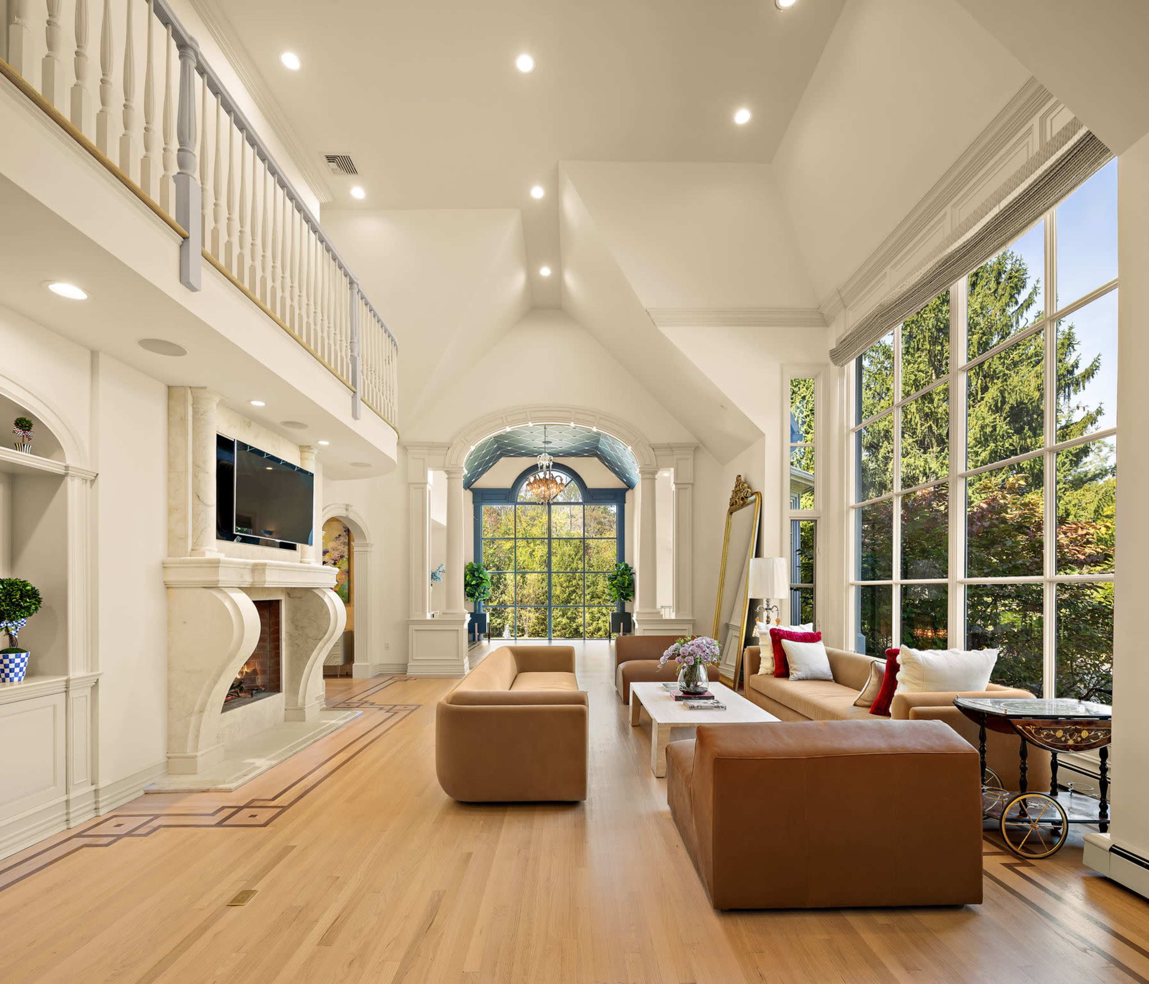 The image shows a spacious living room featuring a high ceiling, large windows, and a fireplace, with modern furniture arranged around a central coffee table.