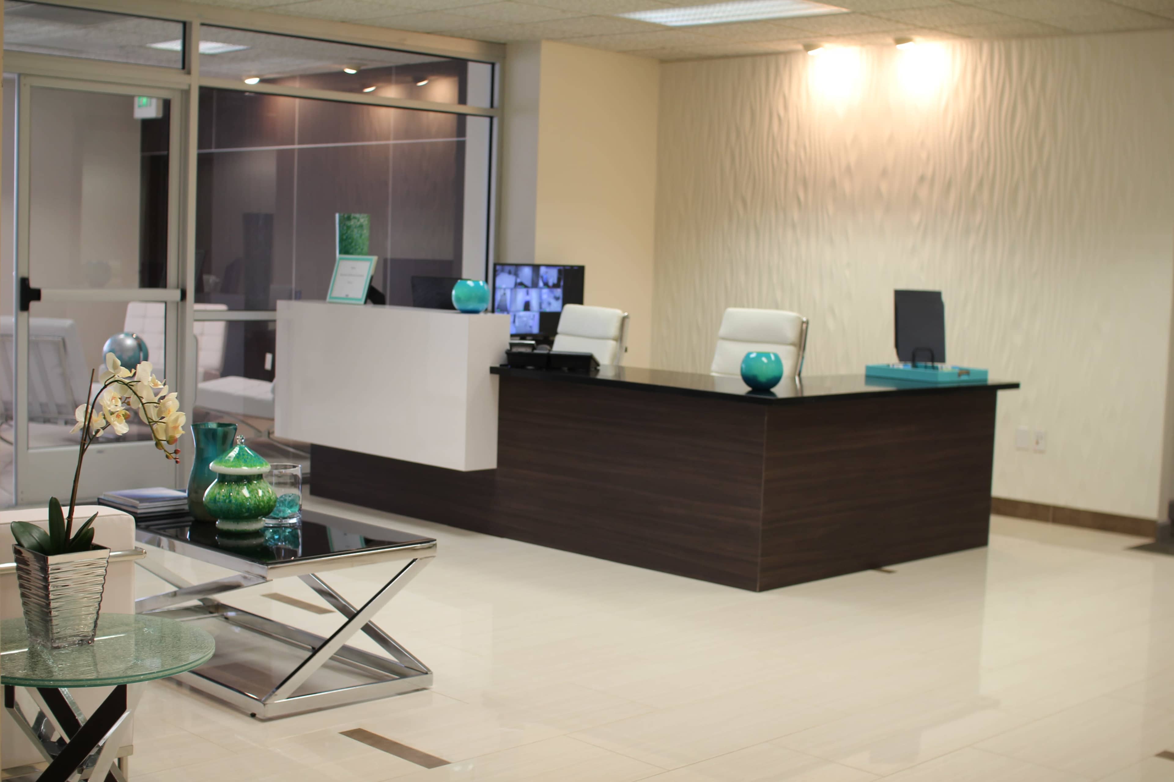 A modern reception area featuring a sleek wooden desk, two white chairs, and decorative elements like plants and turquoise accessories.