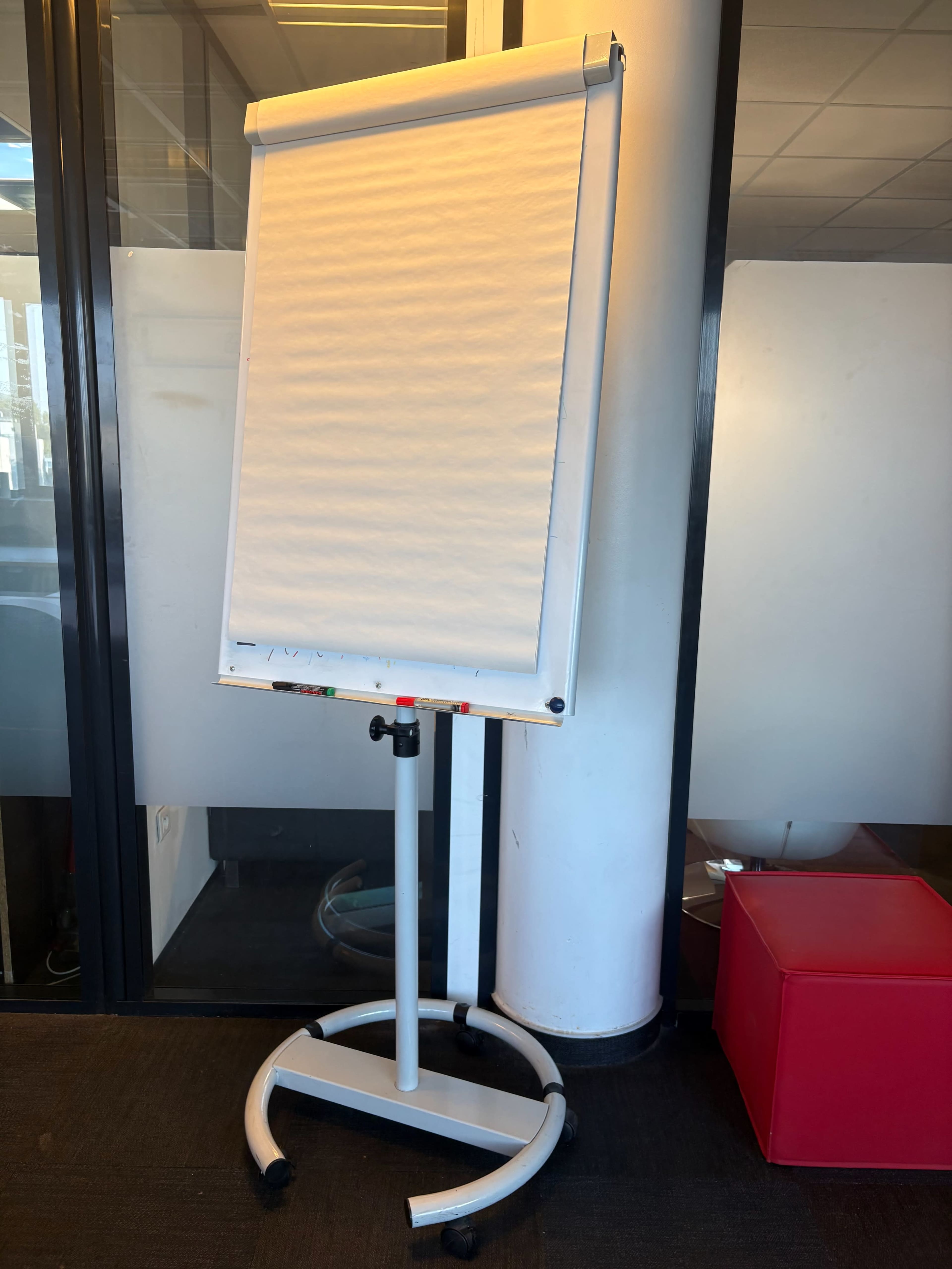 A blank flip chart stand is positioned next to a glass wall in an office setting.