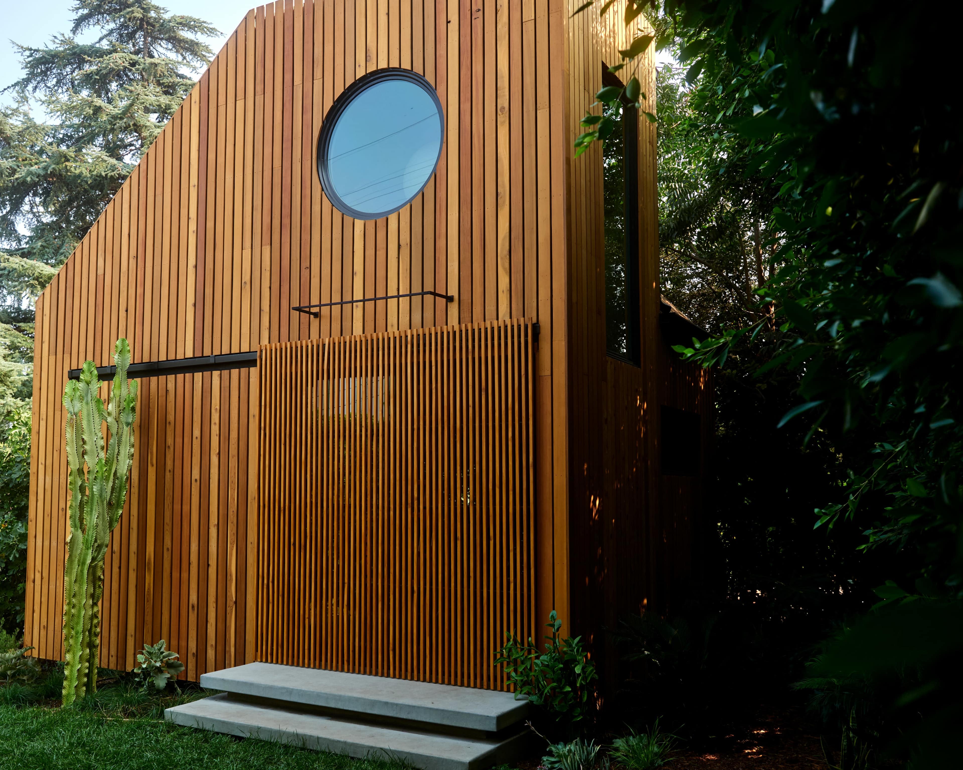 A wooden structure with a circular window features vertical slats and is surrounded by greenery in a garden setting.