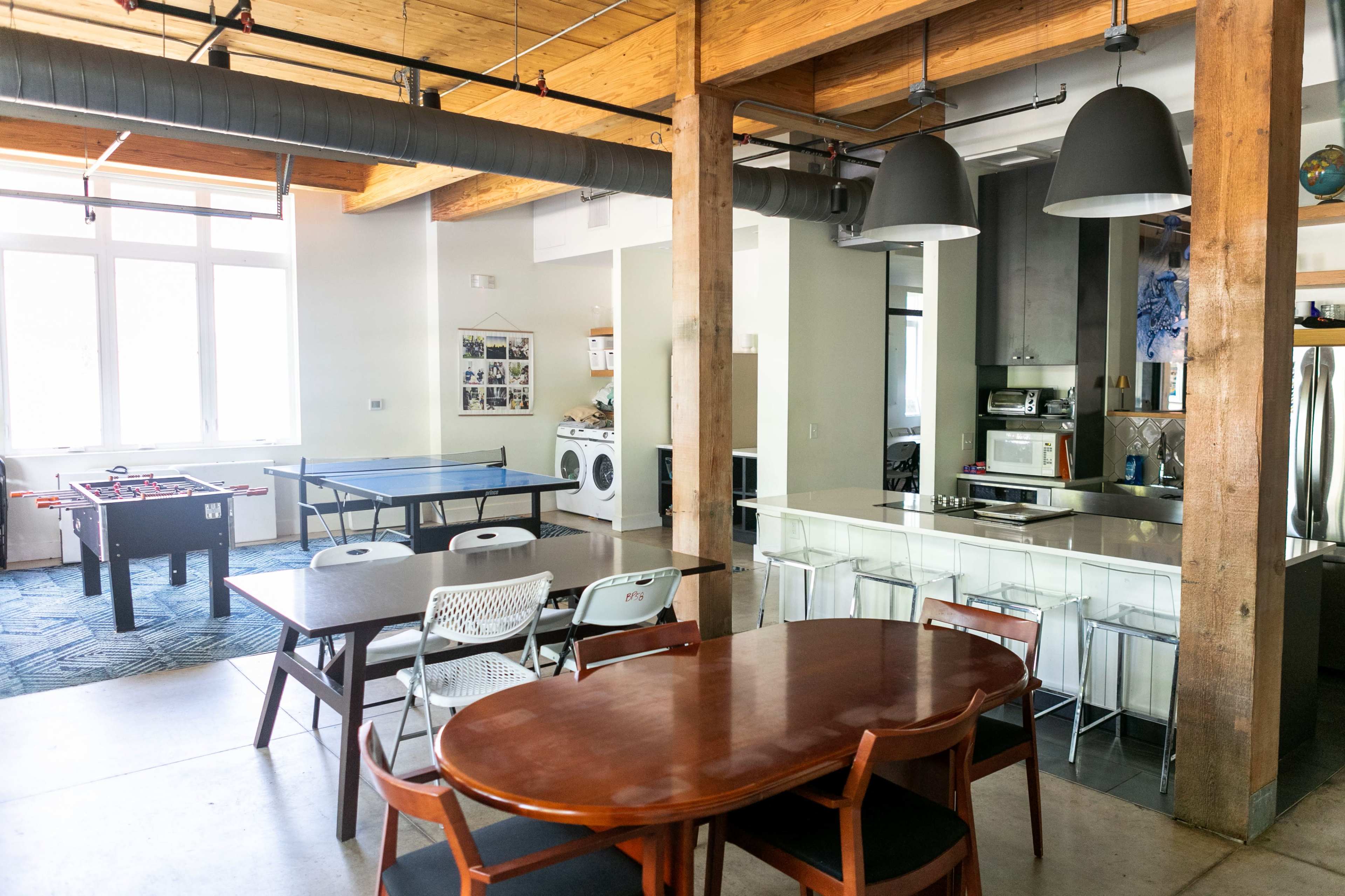 The image shows a modern communal space featuring a kitchen area, dining tables, a game area with a foosball table and a ping pong table, all illuminated by natural light.