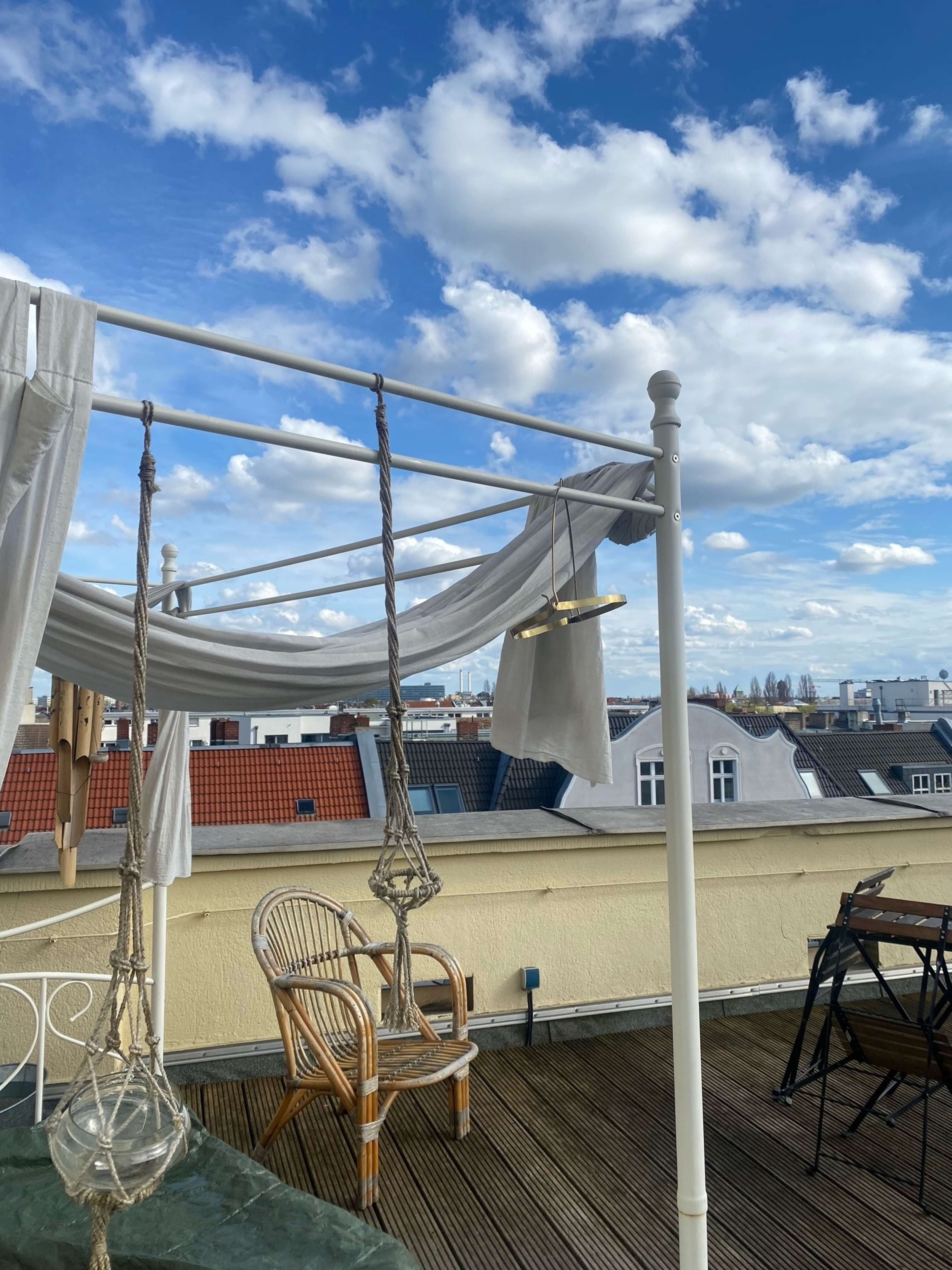 A rooftop terrace with a wooden deck, a hanging swing chair, and white fabric draped over a frame under a partly cloudy sky.
