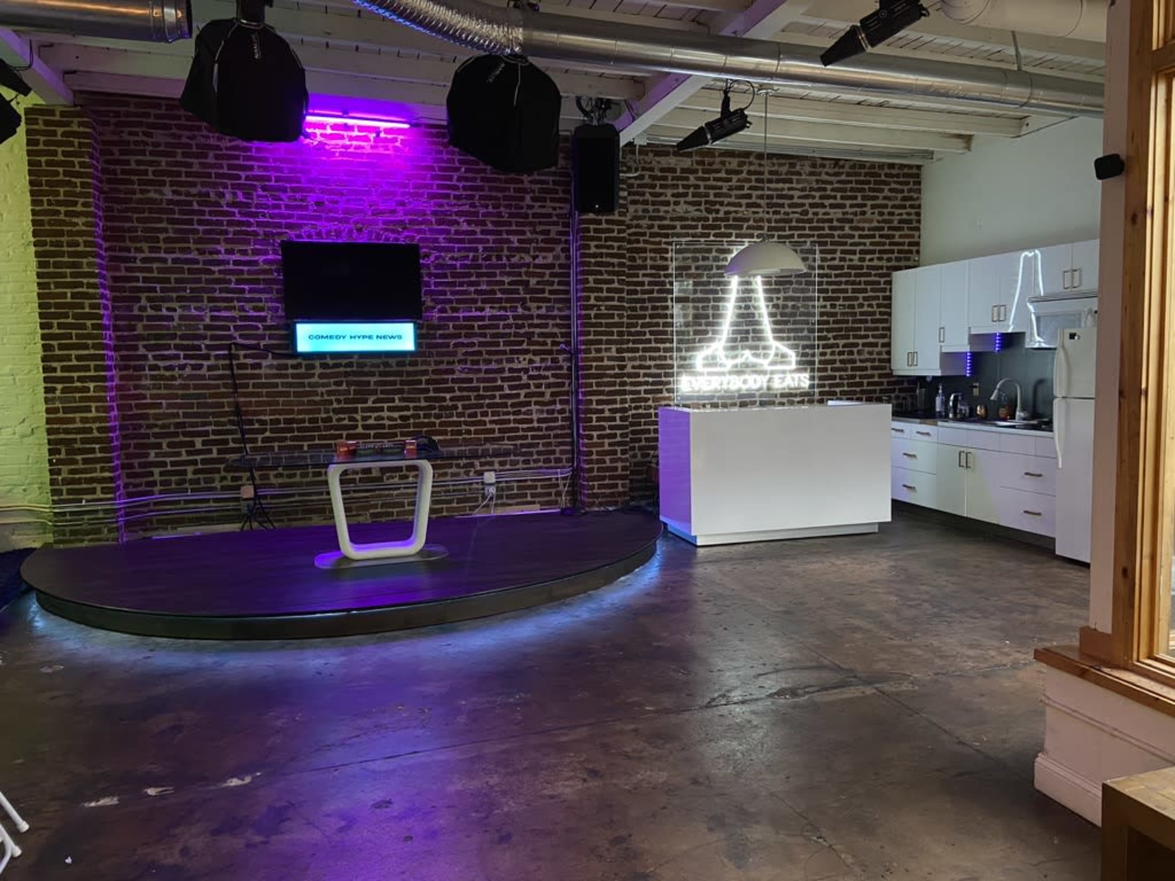 A modern interior space with exposed brick walls, featuring a circular stage area, a kitchen with white cabinetry, and neon lighting accents.