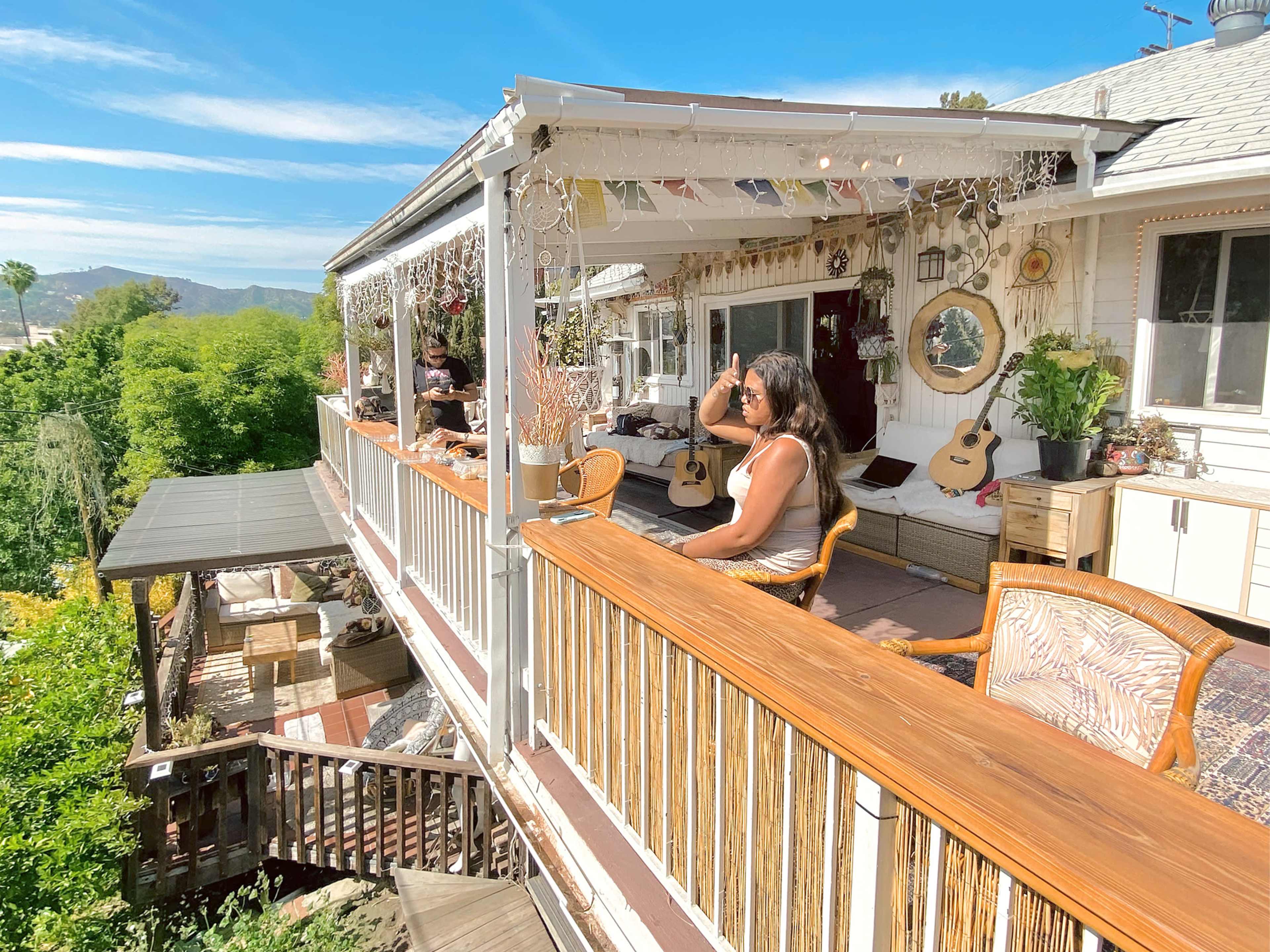 A woman sits on a deck with wooden railings, surrounded by plants, guitars, and decorations, overlooking a scenic view.
