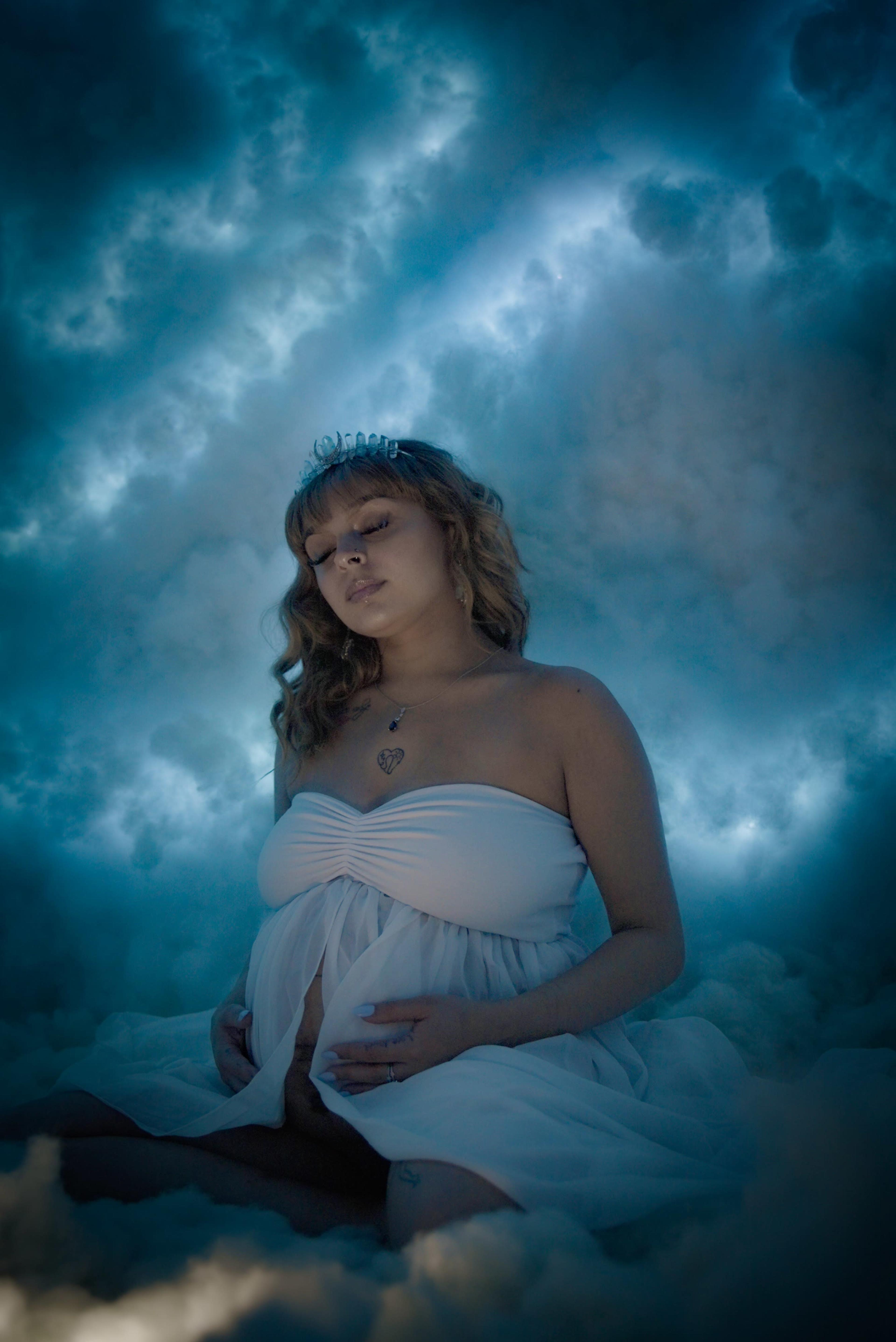 A woman in a white dress sits against a backdrop of blue-tinted clouds, gently cradling her abdomen.