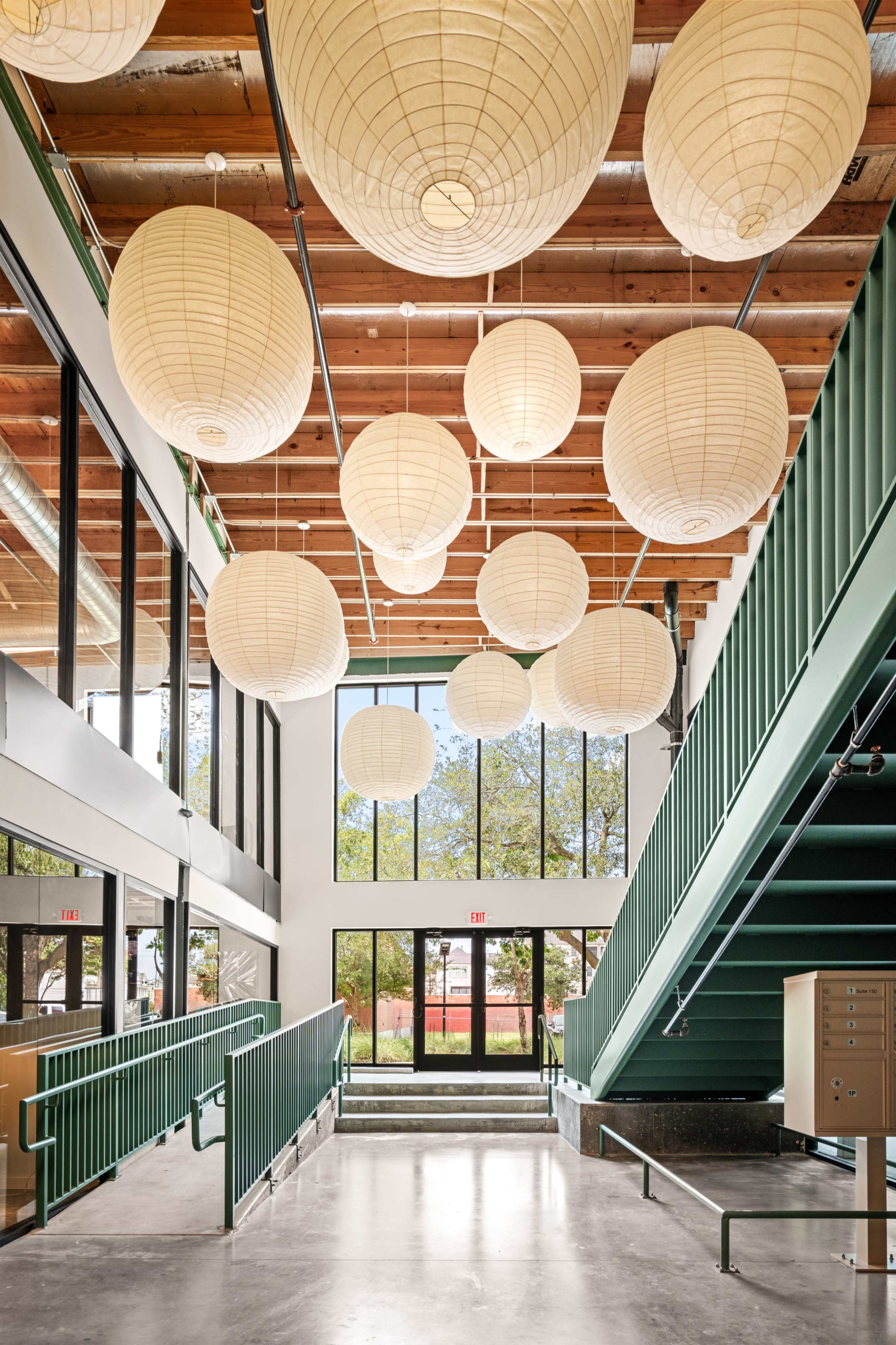 The interior features a spacious lobby with large paper lanterns hanging from the ceiling, a green staircase, and large windows allowing natural light to enter.