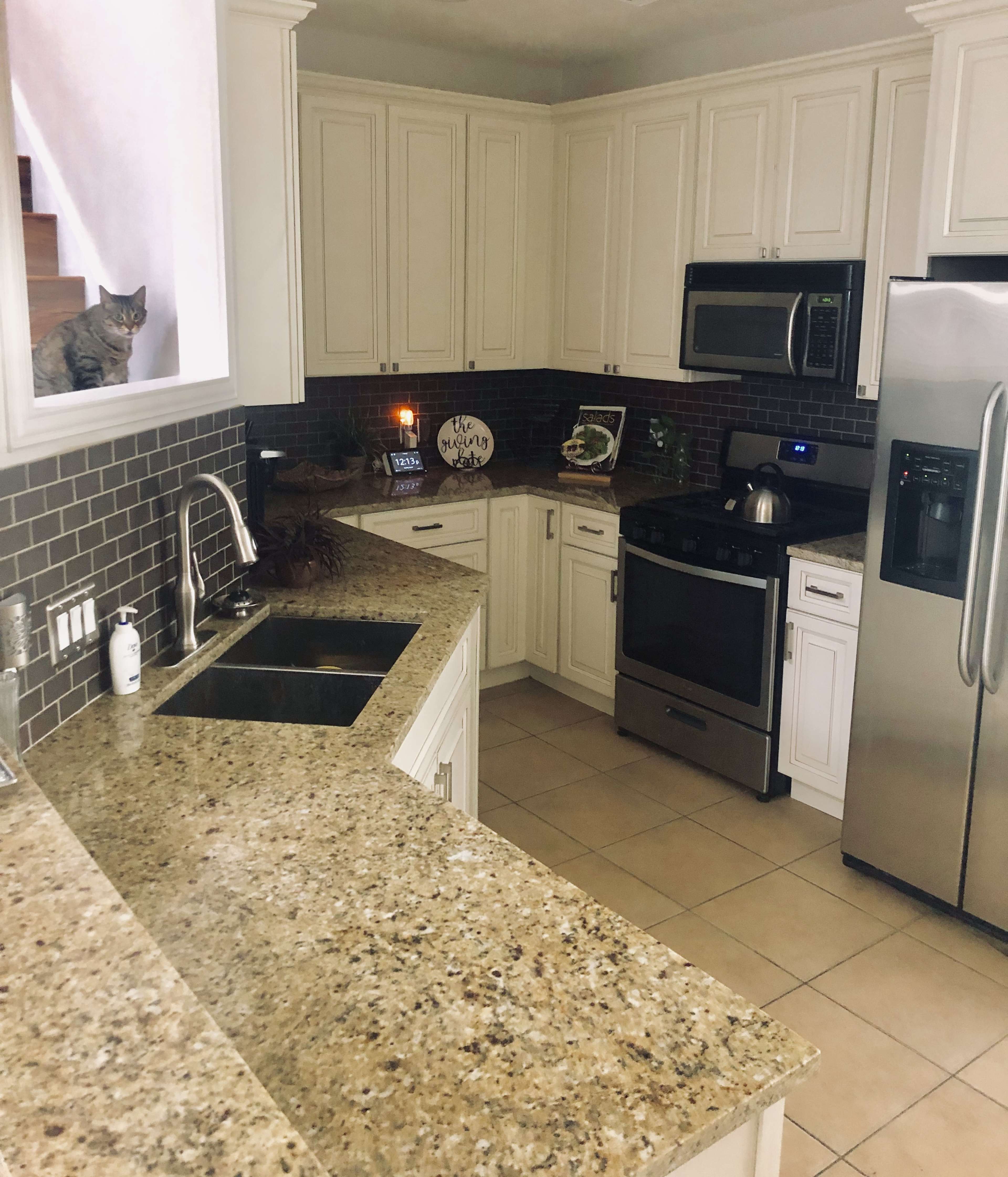A modern kitchen features granite countertops, stainless steel appliances, and a cat sitting on the windowsill.