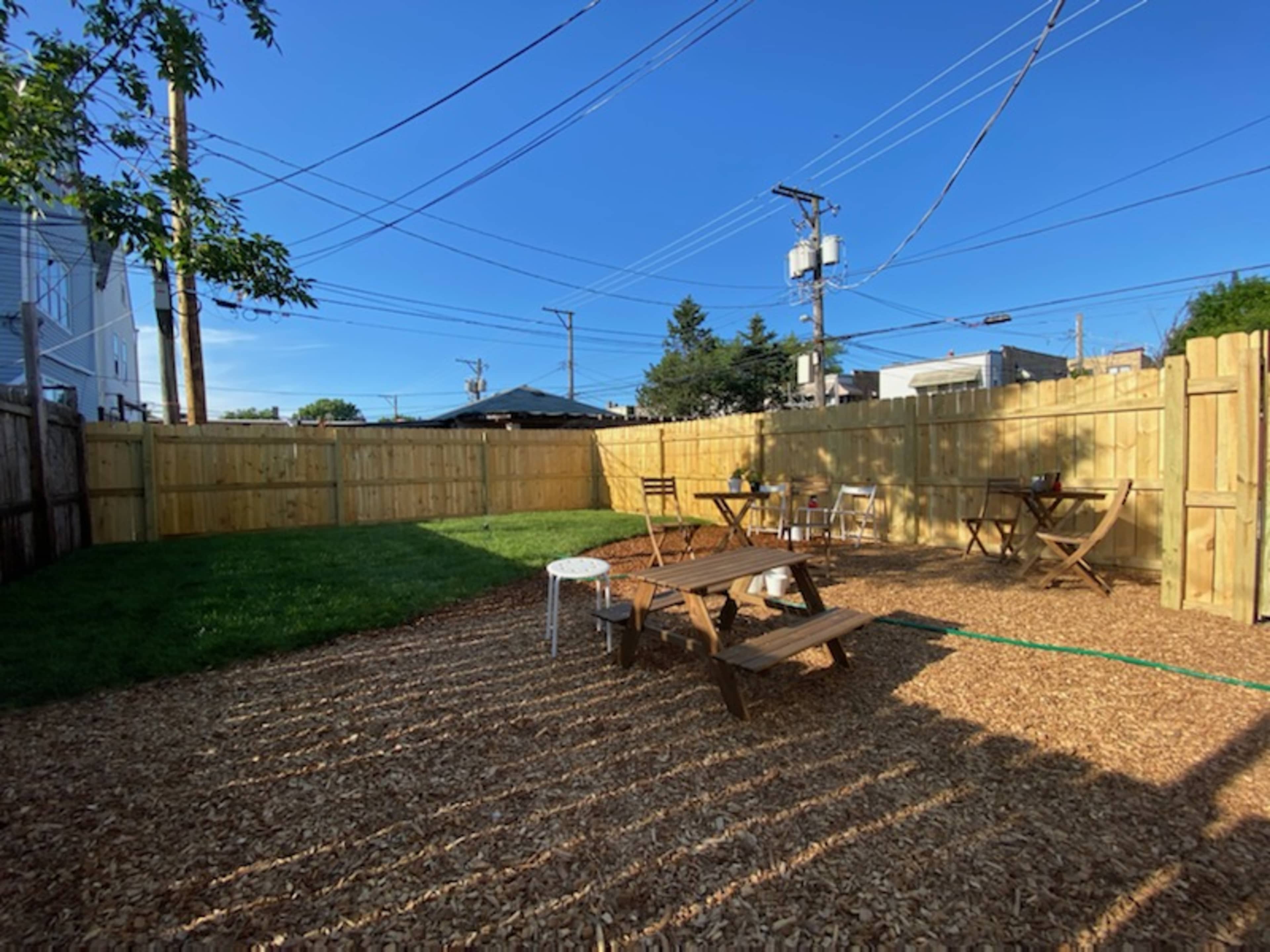 The image shows a fenced outdoor space with wooden seating, gravel, and grass under a clear blue sky.