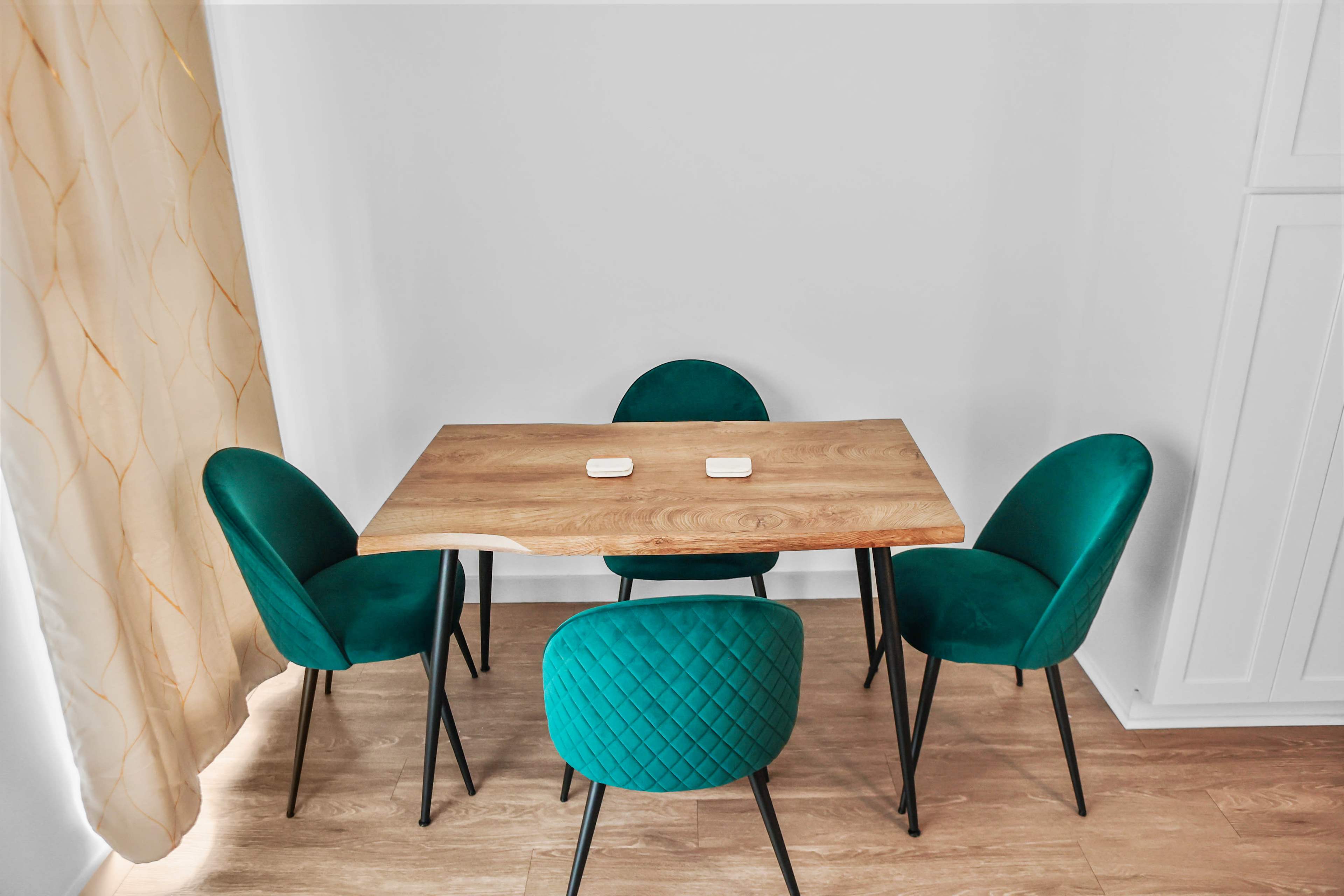 A wooden table with two white coasters is surrounded by four green upholstered chairs in a bright room with a light curtain.