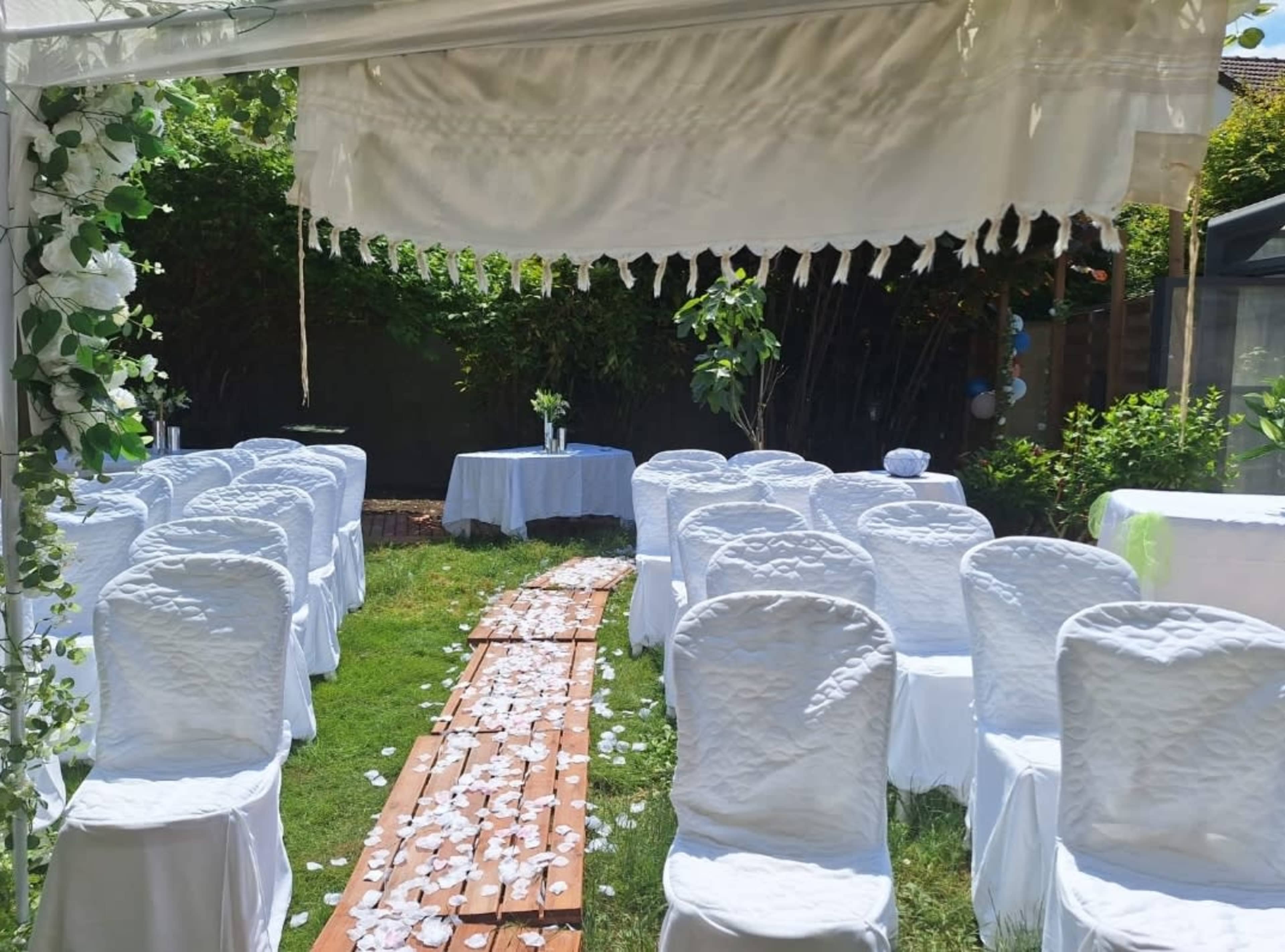 The image shows a wedding setup with white chairs arranged in rows on grass, a wooden pathway covered in flower petals, and a small decorated table in the background.