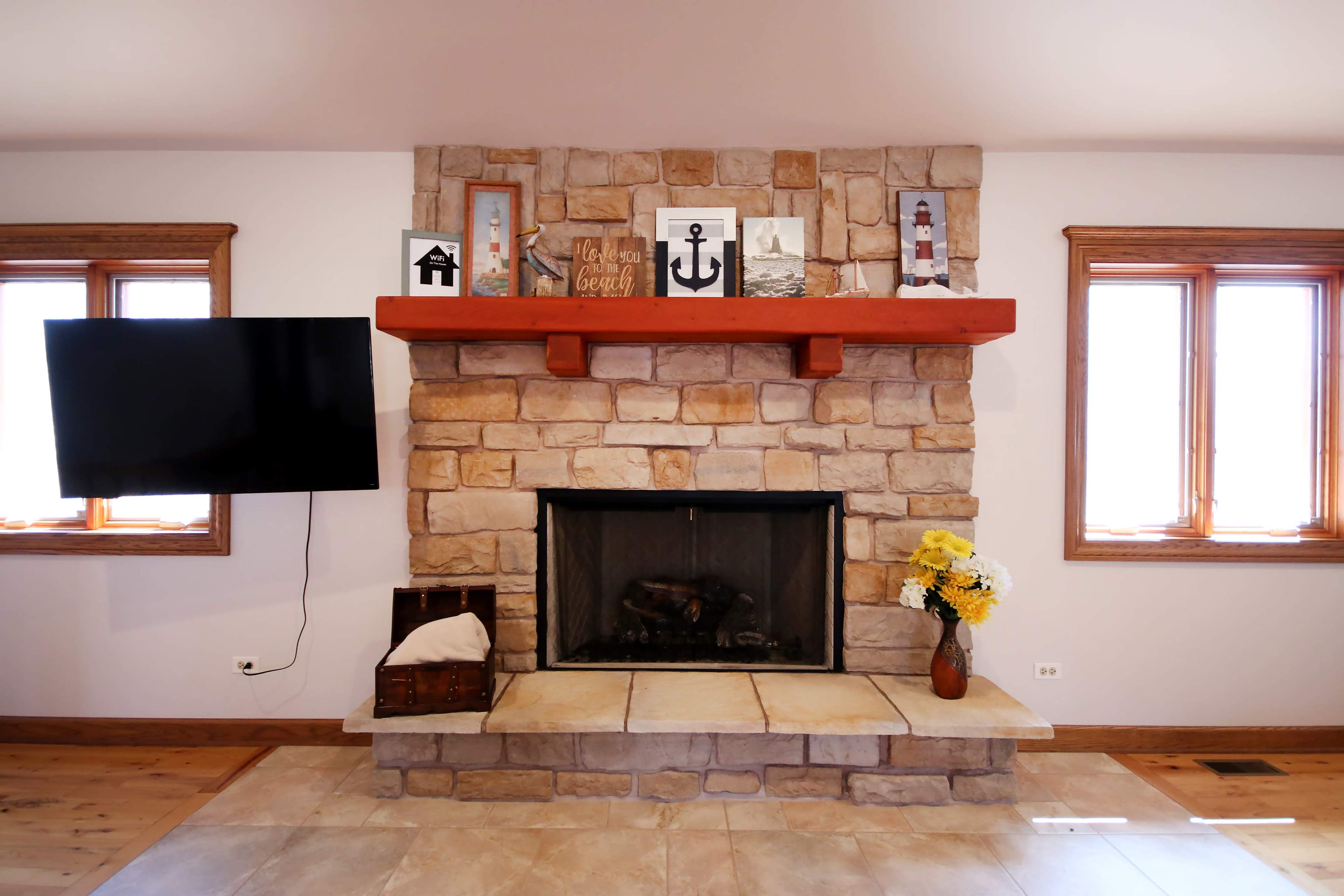 A stone fireplace with a wooden mantel, a television mounted on the wall, and two windows on either side, adorned with a decorative arrangement above the mantel.