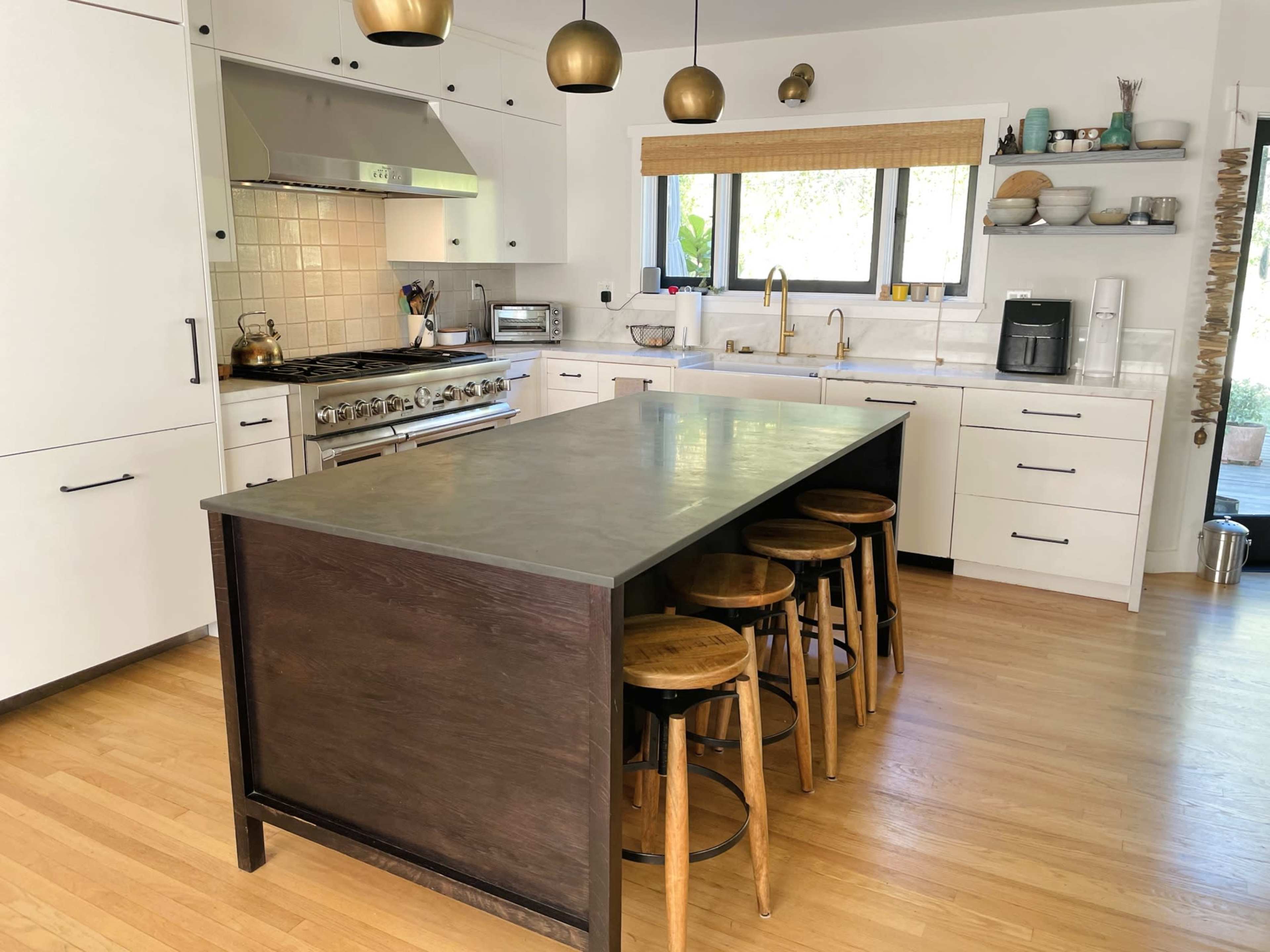 A modern kitchen features a central island with wooden stools, stainless steel appliances, and a large window allowing natural light to enter.
