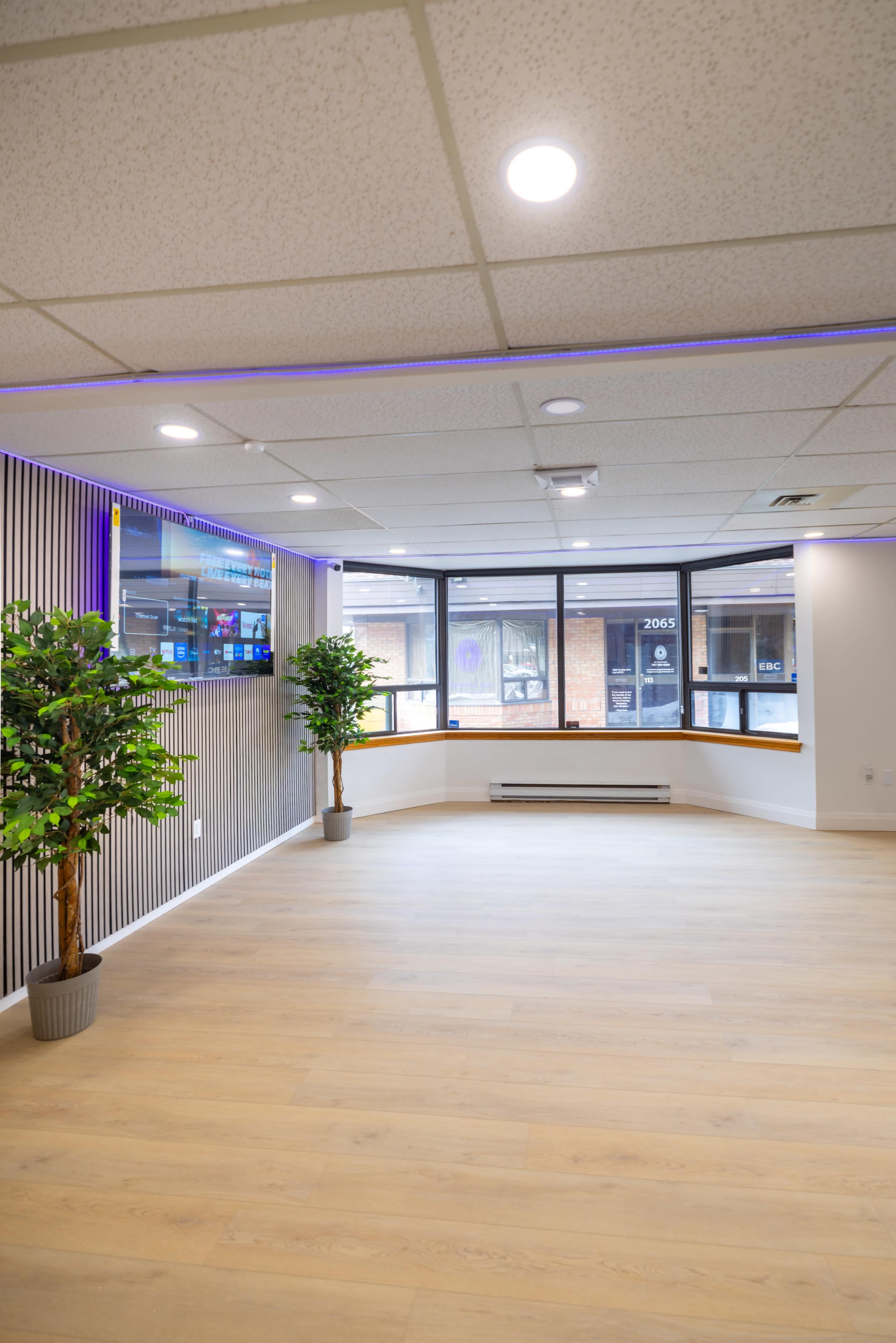 The image shows a spacious, modern interior with wooden flooring, two potted plants, ambient lighting, and large windows facing a street.