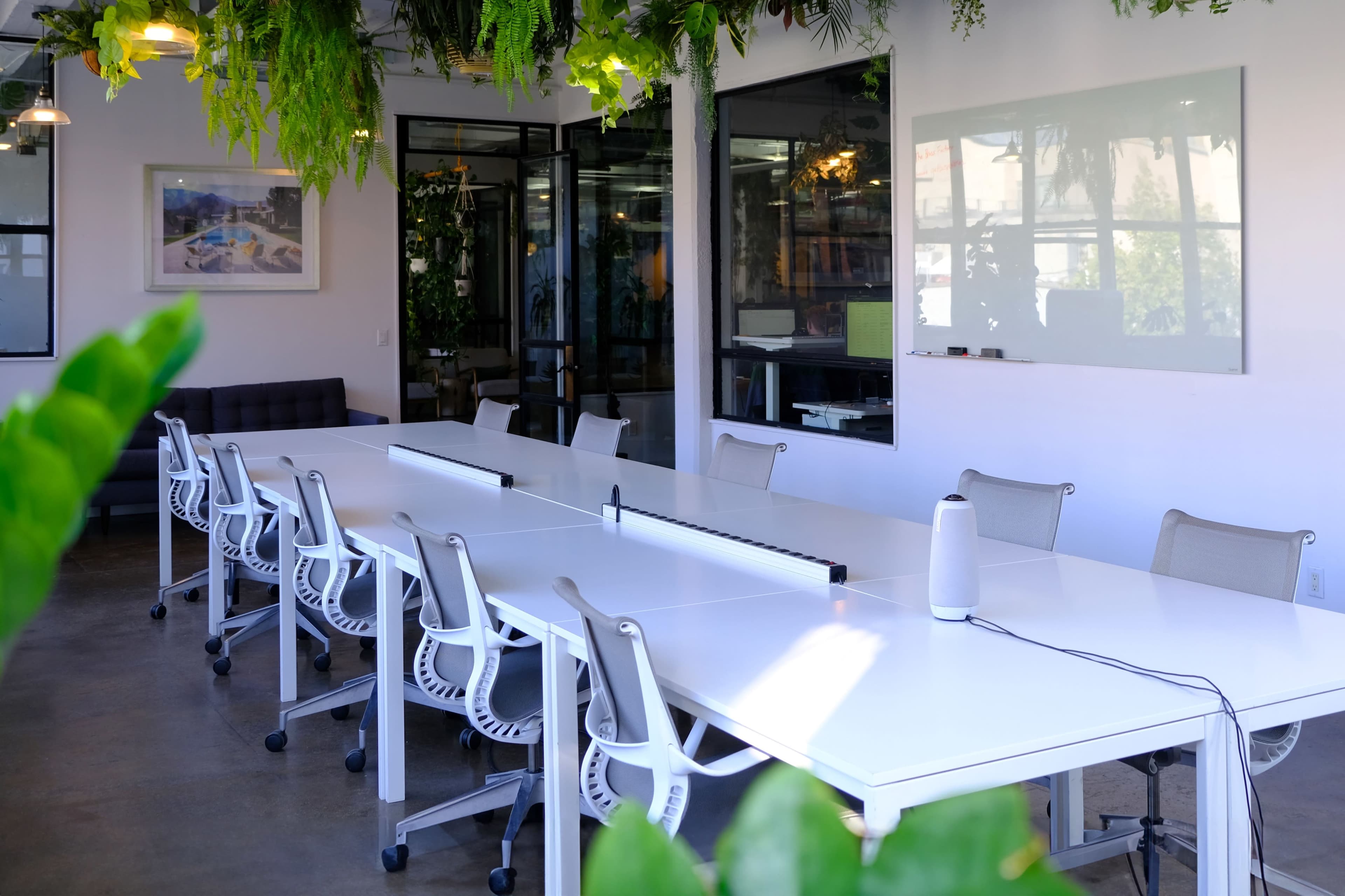 A long, white conference table with several ergonomic chairs is set in a brightly lit room adorned with hanging plants and large glass doors.