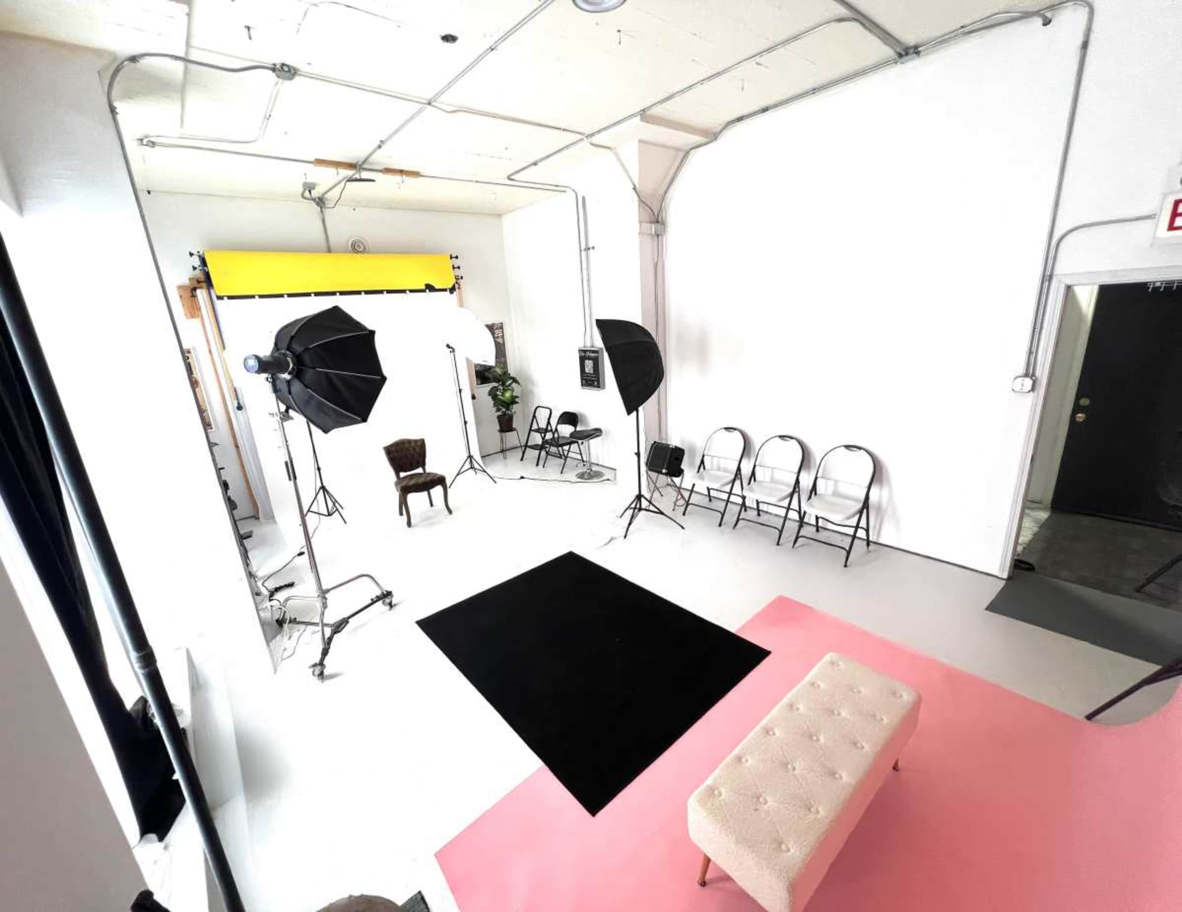 The image shows a photography studio with a plain backdrop, lighting equipment, a chair, and folding chairs arranged around the space.