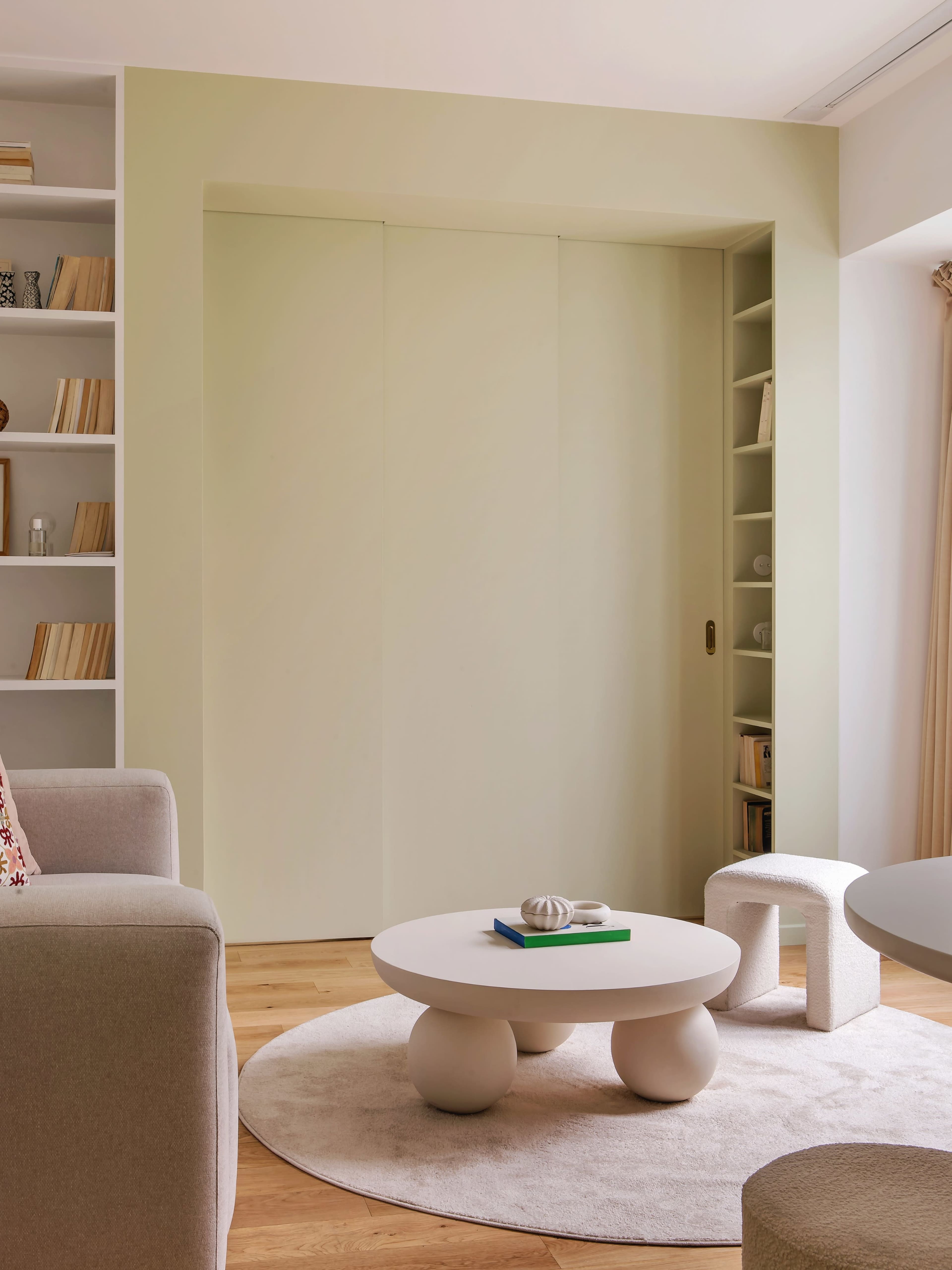 The image shows a minimalist living area with light-colored walls, a round coffee table, a small stool, a plush sofa, and a built-in bookshelf.
